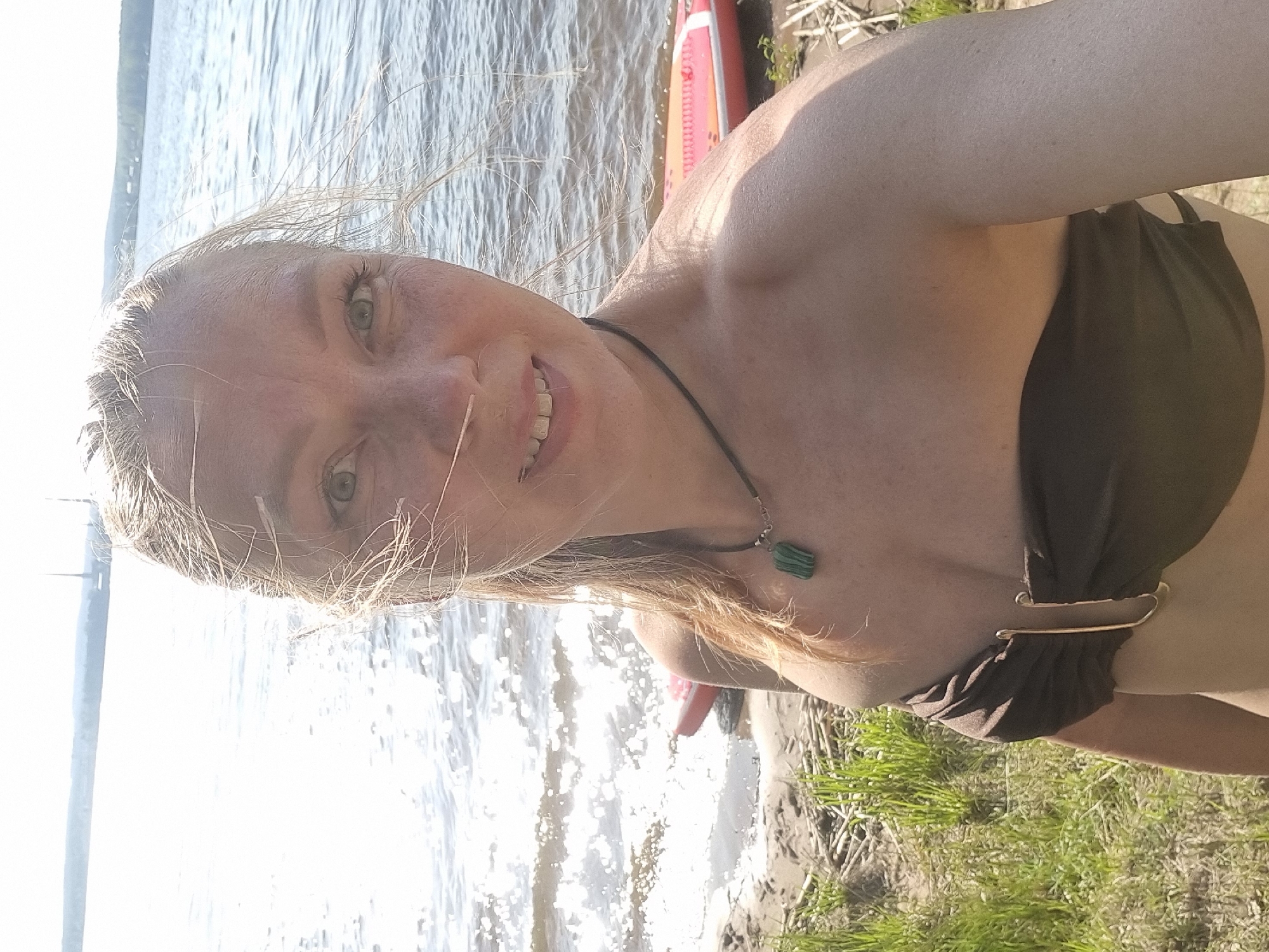 A woman stands on a bank, presumably near water. She is facing the camera, smiling. She is wearing a strapless top and a necklace with a green pendant. A red kayak is partially visible in the background. The scene includes water, grass, and dirt. The location is identified as the city of Chaykovsky, Russia.