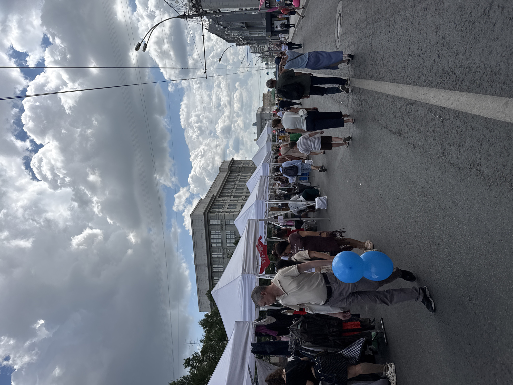 The image depicts an outdoor market scene on a street in Novosibirsk, Russia, under a cloudy sky. Tents are set up along the street, displaying various items, likely clothing, for sale. Several people are browsing the stalls or walking down the street, some carrying bags. In the foreground, a man is walking while holding two blue balloons. The buildings visible in the background are typical of urban architecture. The overall impression is that of a lively market day in the city.
