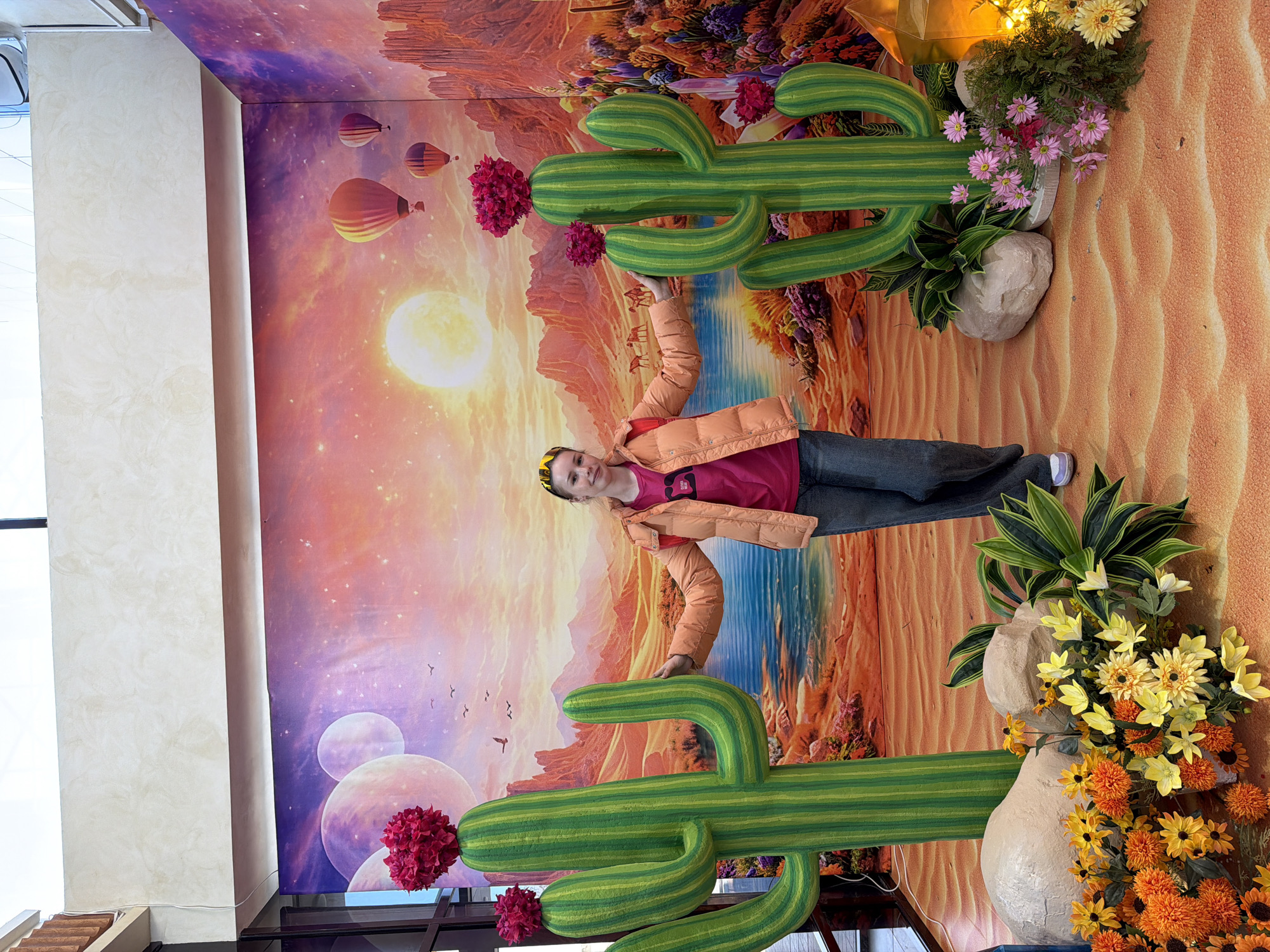 A woman stands in front of a vibrant, surreal mural. The mural depicts a desert landscape with a bright sun, hot air balloons, and fantastical planets. Large, artificial cacti with flowers are positioned in front of the mural. The foreground is decorated with rocks and colorful artificial flowers. The woman wears a pink shirt, orange jacket, and gray pants. The location is Berezniki, Russia.