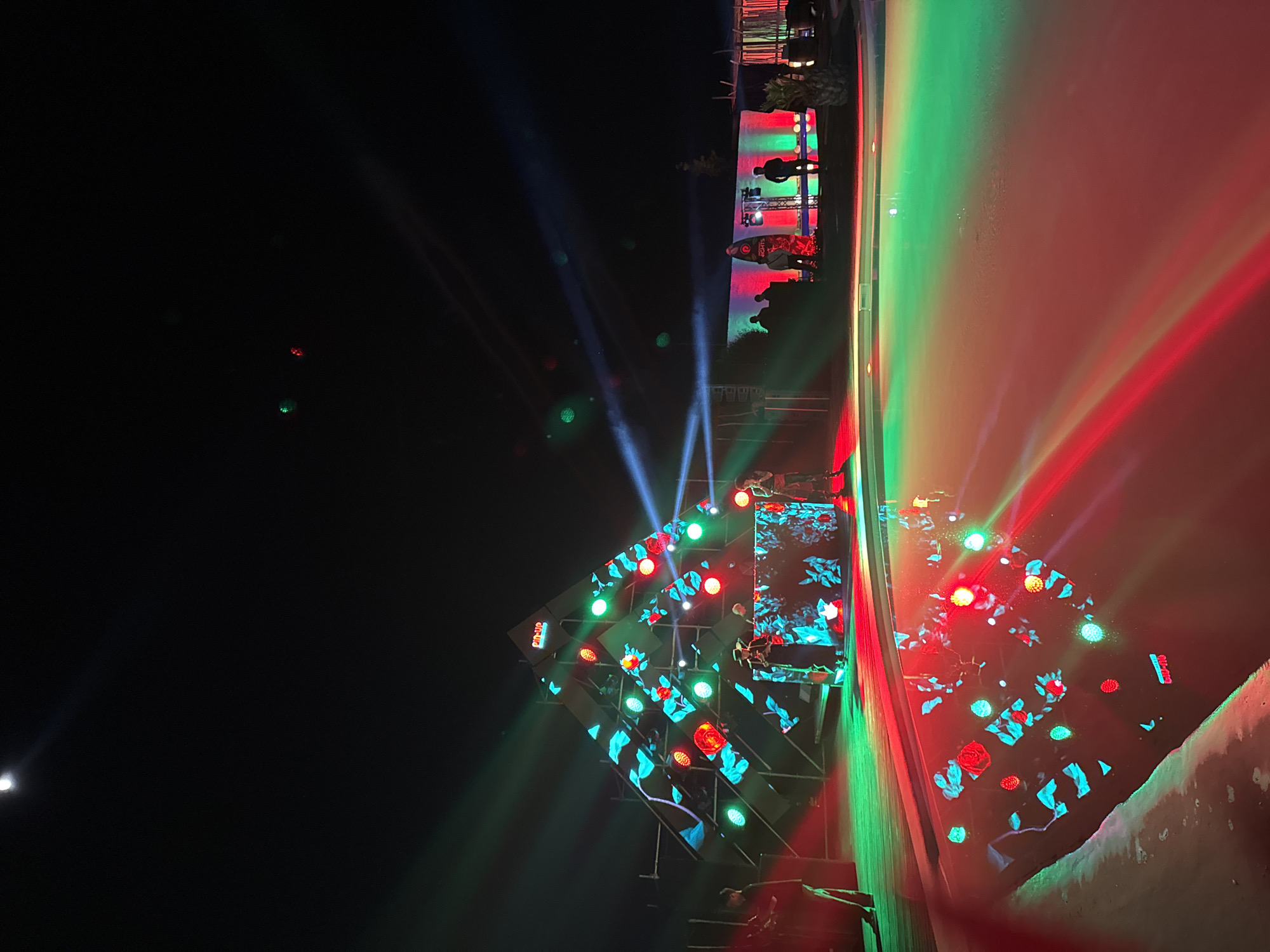 This image captures an outdoor night event in Parakar, Armenia, featuring a vibrant light show and a stage setup. The scene is dominated by a stage structure illuminated with dynamic, colorful lights, primarily red, green, and blue.
The main subject is a large, multi-panel LED screen structure forming a geometric shape, possibly a pyramid or prism. These screens display animated patterns, including what appear to be red roses and teal-colored leaves. Scattered across and around the stage structure are numerous circular spotlights, glowing in red and green. Bright blue, red, and green light beams crisscross the dark sky, emanating from the stage area.
In the background, on a slightly elevated platform or balcony to the right of the stage, several silhouetted figures of people are visible, some standing near a railing, observing the performance. Beyond this platform, lush green plants add a touch of natural element. A long, light-colored wall runs along the right side of the frame, reflecting intense red and green light from the stage, creating a striking color gradient. A bright white light source, possibly the moon or a powerful floodlight, is visible in the upper left of the dark sky.
Visible text includes "NIGHTS" on a banner near the audience area, and "ON-AIR" and "CUPID" on different sections of the LED stage structure. The overall atmosphere is festive and lively, indicative of a concert, festival, or a similar performance event.