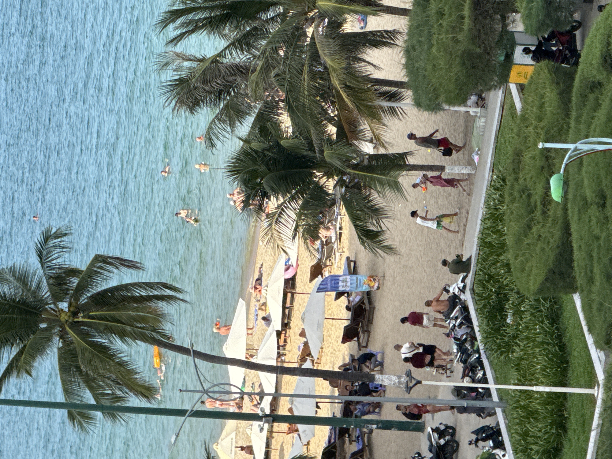 The image depicts a bustling beach scene in Nha Trang, Vietnam, on a bright, clear day. The turquoise ocean stretches to the horizon, dotted with numerous swimmers and a yellow inflatable raft. The sandy beach is lined with white umbrellas and lounge chairs, many occupied by sunbathers. Palm trees, characteristic of the tropical climate, provide shade and a sense of place.
In the foreground, a paved walkway separates the beach from a lush green area with neatly trimmed hedges and a lamppost. Several motorcycles are parked along the walkway, indicating local transportation. People are visible on the beach, some walking along the shore, others relaxing under umbrellas, and a few are playing in the shallow water. A group of individuals are gathered near the parked motorcycles, appearing to be either arriving or departing. The overall atmosphere is one of leisurely activity and vibrant coastal life.