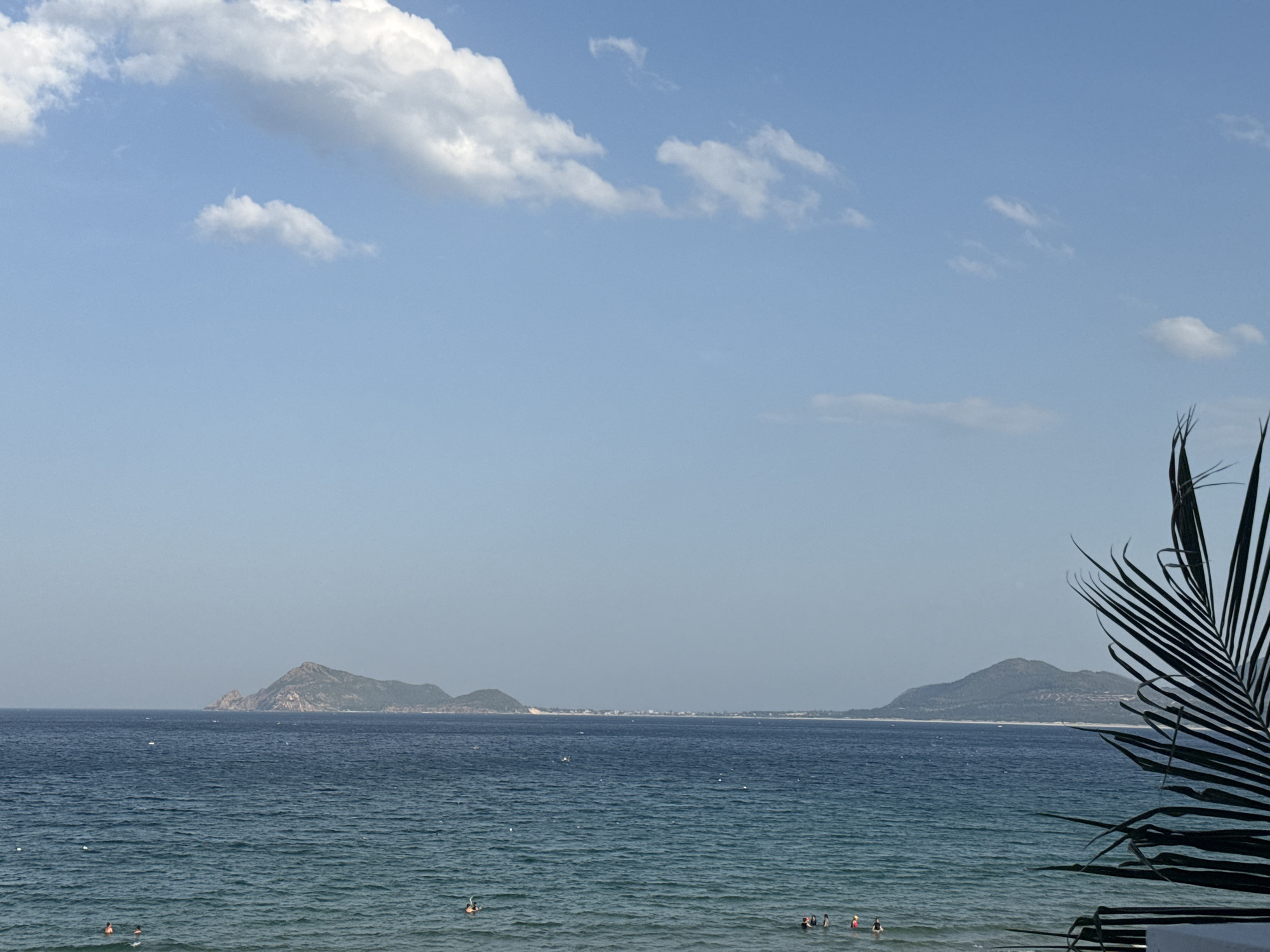 The image depicts a tranquil scene at a beach in Unknown, Unknown. The sky is a clear, bright blue, dotted with a few fluffy white clouds, most notably a large bank of clouds in the upper left quadrant and a few smaller, wispy clouds scattered across the expanse. The sea is a deep, inviting blue, with gentle ripples indicating a calm day. In the distance, several islands with rounded, green-topped hills rise from the horizon. A faint, hazy line of coastline with buildings can be observed between the islands. In the foreground, a few people are visible enjoying the water; some are swimming, while others appear to be wading. The right side of the image is framed by the dark, silhouetted fronds of a palm tree, adding a tropical touch to the scenery.