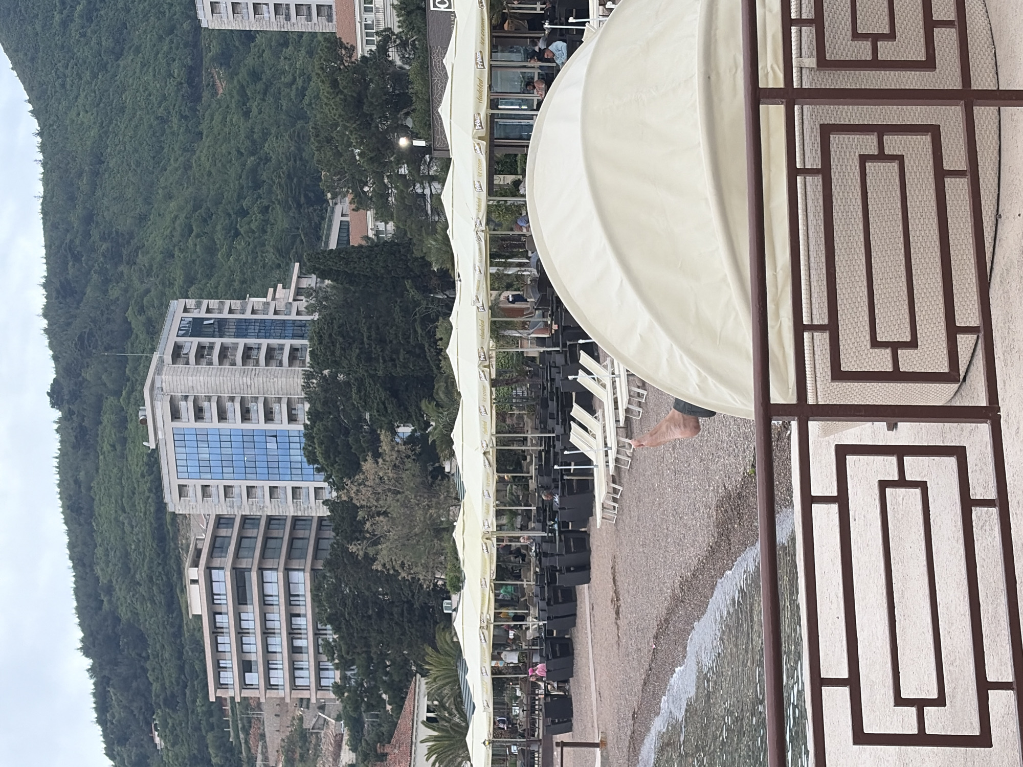 The image shows a beachside scene in Budva, Montenegro. A building with numerous balconies and windows is positioned on a hillside covered with dense trees. On the beach, there is a row of umbrellas and chairs, and a restaurant appears to be in operation. A large, white cabana-like structure with brown metal rectangular frames dominates the foreground of the image. Water is lapping at the pebbly beach. The sky is overcast. People are seated at some of the tables. A bare foot is visible.