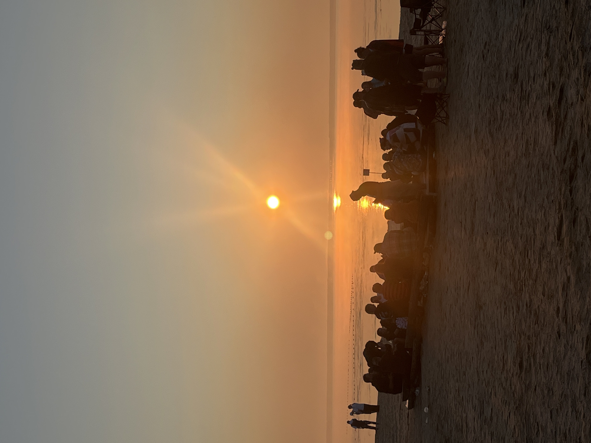 A group of people are gathered on a sandy beach in Surakovo, Russia, facing a body of water during sunset. The sun is low on the horizon, casting a warm orange glow across the sky and water, with visible sun rays. Some individuals are seated on wooden benches, while others stand. Two portable chairs are visible on the right side of the frame. The scene appears to be a social gathering or event taking place outdoors.
