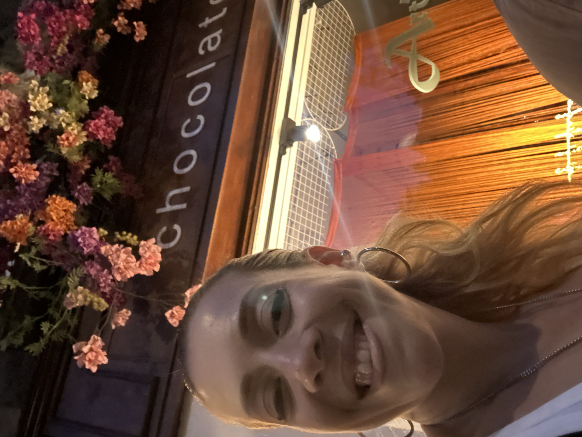 A woman takes a selfie in front of a shop with "Chocolat" written in white lettering. The shop appears to be decorated with colorful flowers above the signage. Behind the woman, the shop window has orange drapes and a white letter "A" visible. The scene is indoors or in a covered area, possibly a street in Amsterdam, Netherlands.