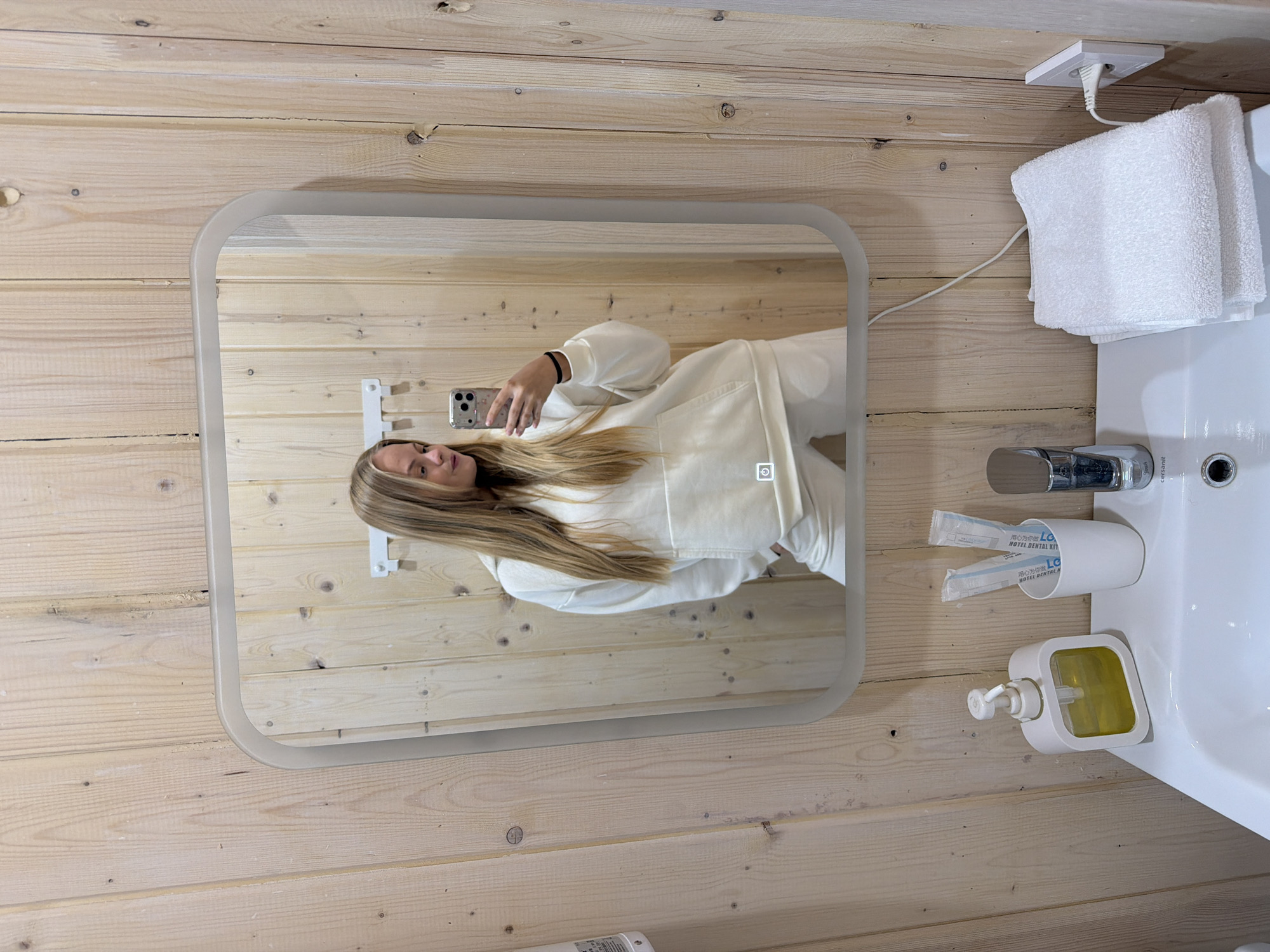 The image captures a selfie taken by a young woman in a modern bathroom, likely within a hotel or guesthouse in Vozhoiskii, Russia. The room's walls are clad in light-colored wooden planks, featuring visible grain and knots, which impart a natural and rustic ambiance.
Reflected in a rectangular mirror with rounded corners, the woman, sporting long, straight blonde hair, is seen holding a smartphone, seemingly an iPhone, to take the picture. She is wearing a cream-colored long-sleeved top or robe and has a subtle smile on her face. The mirror itself is illuminated, featuring a frosted edge that emits light, and a touch-sensitive power icon is visible on its surface, indicating a light function.
To the right of the mirror, a white ceramic sink is equipped with a contemporary chrome faucet. A neatly folded white towel rests above the sink, beneath a white electrical outlet where a cord is plugged in, extending towards the mirror, presumably powering its illumination. Adjacent to the faucet, a white cylindrical holder contains two individual packages labeled "HOTEL DENTAL KIT" with additional text in Chinese: "用心为你做 LG". Below this, a square-shaped white pump dispenser filled with yellow liquid, likely soap, is positioned by the sink. The brand "cersanit" is faintly visible on the sink itself.
The setting suggests a clean and well-maintained accommodation, captured indoors, with the lighting primarily provided by the mirror's illumination and possibly ambient artificial light.