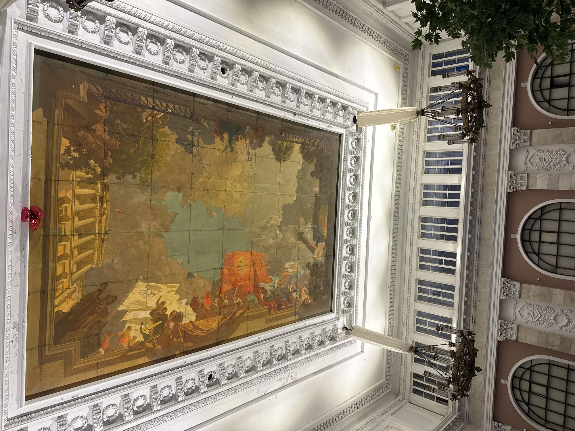 Here is a detailed description of the image:
The photograph captures an ornate interior scene, likely a historical building, from a low-angle perspective looking upwards. The focal point is a large, framed painting inset within the ceiling. The artwork depicts a classical scene with figures in ancient-style clothing on a boat, along with architectural elements and what appears to be an Olympics flag. The painting is bordered by a detailed white frame with classical motifs.
The surrounding ceiling is white and heavily ornamented with molding and decorative trim. Two ornate chandeliers hang down, partially obscured by white fabric or paper covers. To the right of the painting is a series of windows, revealing an exterior wall with additional architectural details, including circular windows. The environment is well-lit, likely by artificial lighting, and the overall impression is of a grand, historic space.
Based on the art style, architecture, and the presence of the Olympics flag, this is likely the Saint Petersburg Polytechnical University, located in Saint Petersburg, Russia. Specifically, it is a portion of the ceiling found inside the University's Main Building.