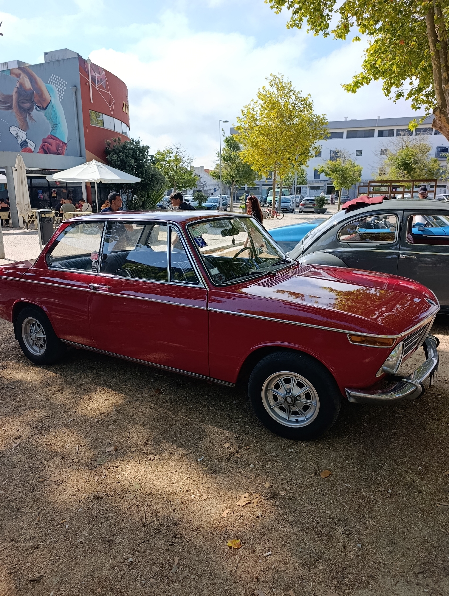 In Setúbal, Portugal, a red vintage BMW 2002 Tii is parked on a gravel surface next to a building with a large mural. The mural depicts a person in mid-air, possibly performing a gymnastic or dance move. The building has a curved facade with geometric patterns and windows. In the background, other vintage cars are visible, including a grey car and a blue car. Several people are present, some seated at tables under white umbrellas outside what appears to be a cafe or restaurant. The scene is illuminated by sunlight filtering through trees, creating dappled shadows on the ground. The sky is partly cloudy.