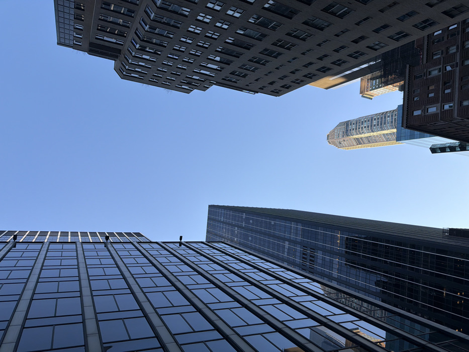 The image displays an upward-looking perspective of multiple skyscrapers and buildings under a clear, light blue sky, indicating bright daylight. The scene is devoid of people, vehicles, or street-level objects, focusing solely on the architecture.
On the far left, a contemporary skyscraper with a dark, reflective glass facade dominates the frame, rising vertically with a grid pattern of rectangular windows. Adjacent to it, slightly further back and to the right, a second, slender, dark glass skyscraper also ascends towards the sky.
In the mid-ground, towards the center-right, a distinctive supertall skyscraper with a lighter-colored, faceted, and crown-like top is prominently featured, illuminated by direct sunlight. This building is identifiable as One57 (157 West 57th Street).
On the far right, a multi-story building with a darker, more traditional brick-like or stone facade, featuring numerous rectangular windows, extends vertically out of the frame. This building is largely in shadow. A shorter, brown building with a glass section is partially visible below it.
The cumulative architectural elements, particularly the presence of One57, definitively place this location within Midtown Manhattan, New York City, United States of America.