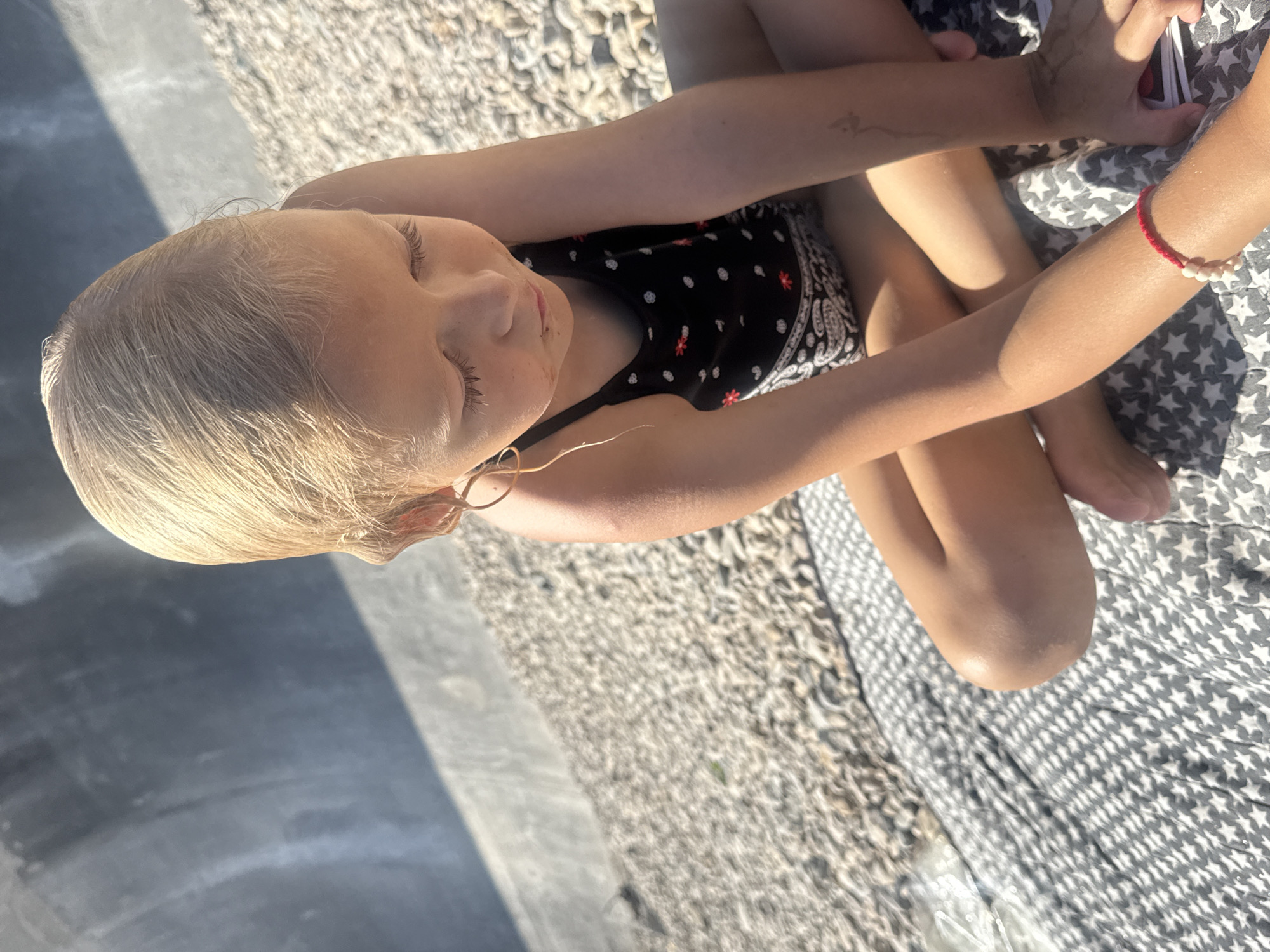 The media file shows a young girl with blonde hair sitting with her eyes closed, her head tilted slightly to the side. She is wearing a black swimsuit with a white and red polka dot pattern. Her arms are resting in her lap, and a red bracelet with white beads is visible on her right wrist. Her legs are crossed. Another person's arm is visible in the upper right corner, holding what appears to be a cell phone or similar device.
The background consists of a textured surface, likely gravel or small stones, and a patch of dark, smooth material that could be a walkway or pavement, with a distinct shadow cast across it. A patterned blanket, possibly with stars, is also visible under the girl.
The lighting suggests it is daytime, possibly with bright sunlight given the shadows. The overall impression is a relaxed, casual setting, possibly outdoors. The girl's closed eyes and relaxed posture convey a sense of peace or rest. There is no visible text in the image.