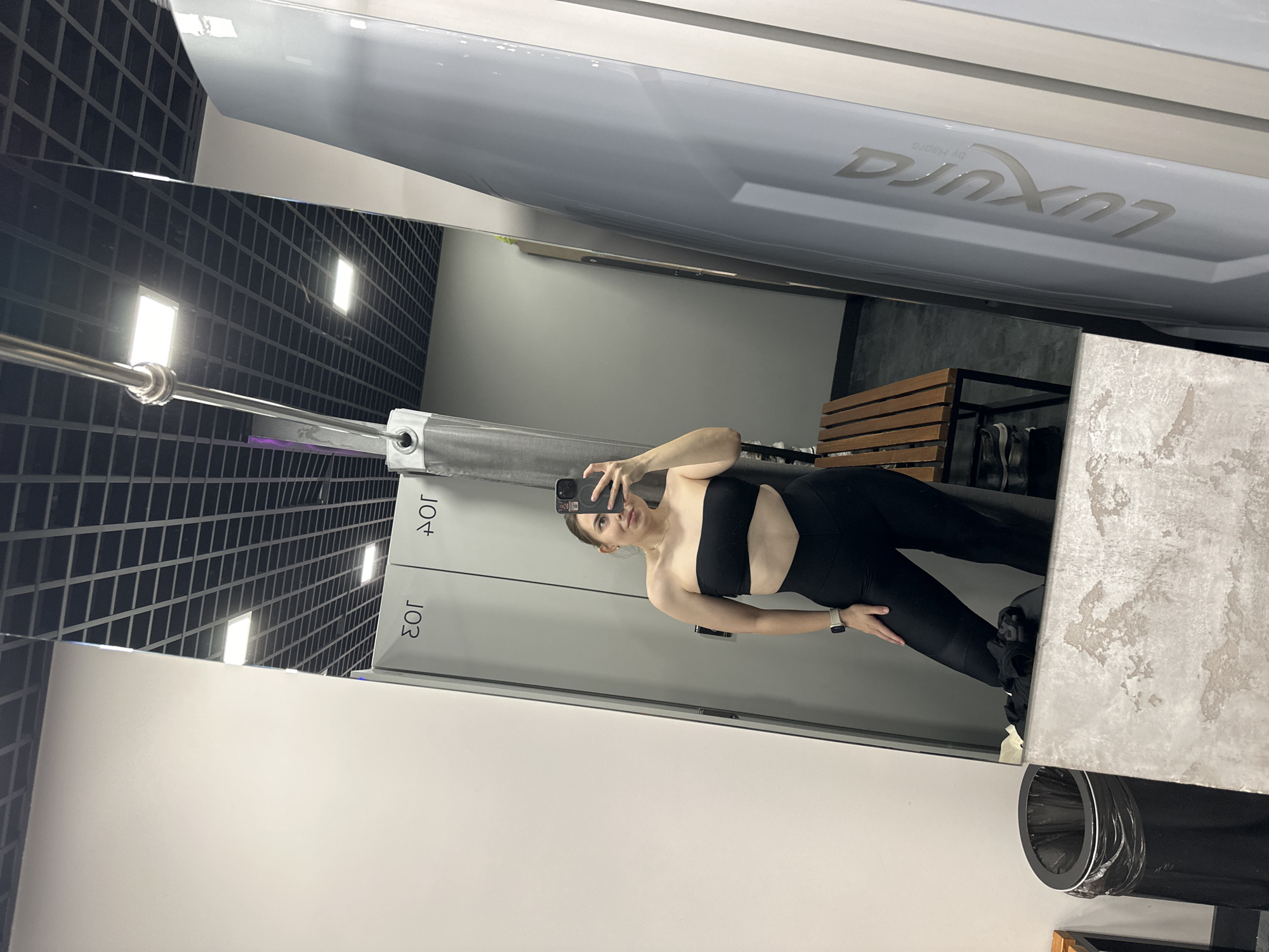 A young woman is taking a selfie in a mirror. She is wearing a black strapless top and black leggings. The mirror is in what appears to be a tanning salon in Perm, Russia, due to the tanning bed reflected in the upper right corner of the mirror and the “JO3” written on the doors. The background includes a black ceiling with a grid pattern, white walls, and a bench. A trash can is visible at the lower right corner.