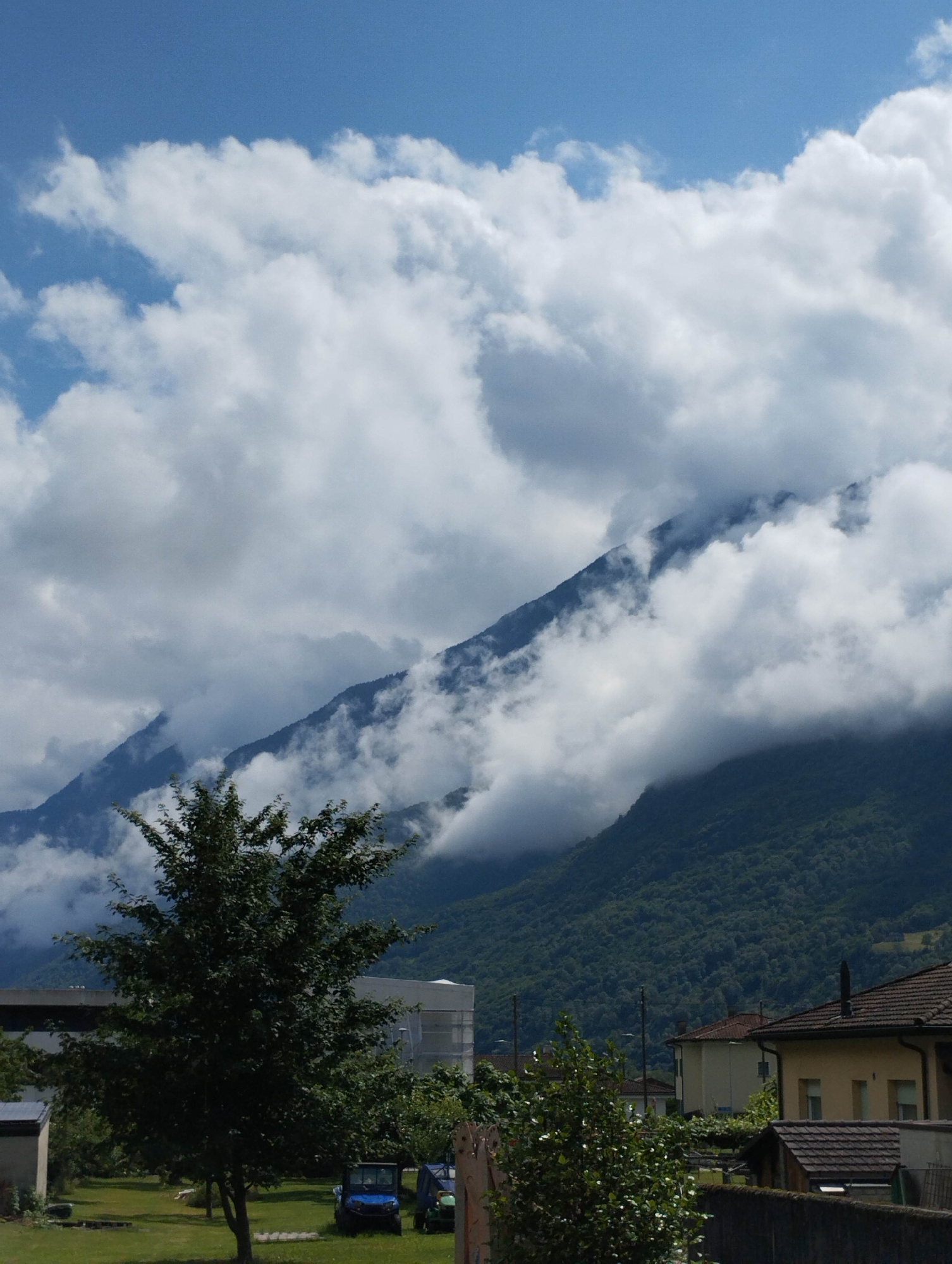 A vertical photograph captures a mountainous landscape under a partially cloudy blue sky in the city of Unknown, Unknown. The upper portion displays a clear blue sky, transitioning to dense white and grey cumulus clouds that obscure the upper and mid-slopes of dark green, heavily forested mountains. Cloud formations are visibly settled among the peaks and valleys of the mountainous terrain. The foreground features a green grassy area. A large, dark green deciduous tree stands centrally in the left foreground. Behind this tree, two utility vehicles, one blue and one green, are parked on the grass. To the left, a light-colored building with a dark roof and a larger, light-grey building, possibly industrial, are partially visible. On the right side of the foreground, residential structures are present, including a two-story house with a yellow facade, brown tiled roof, and a visible chimney. A low, dark wall or fence extends across the bottom right, separating the grassy area from the houses. The scene is illuminated by natural daylight. No individuals are present.