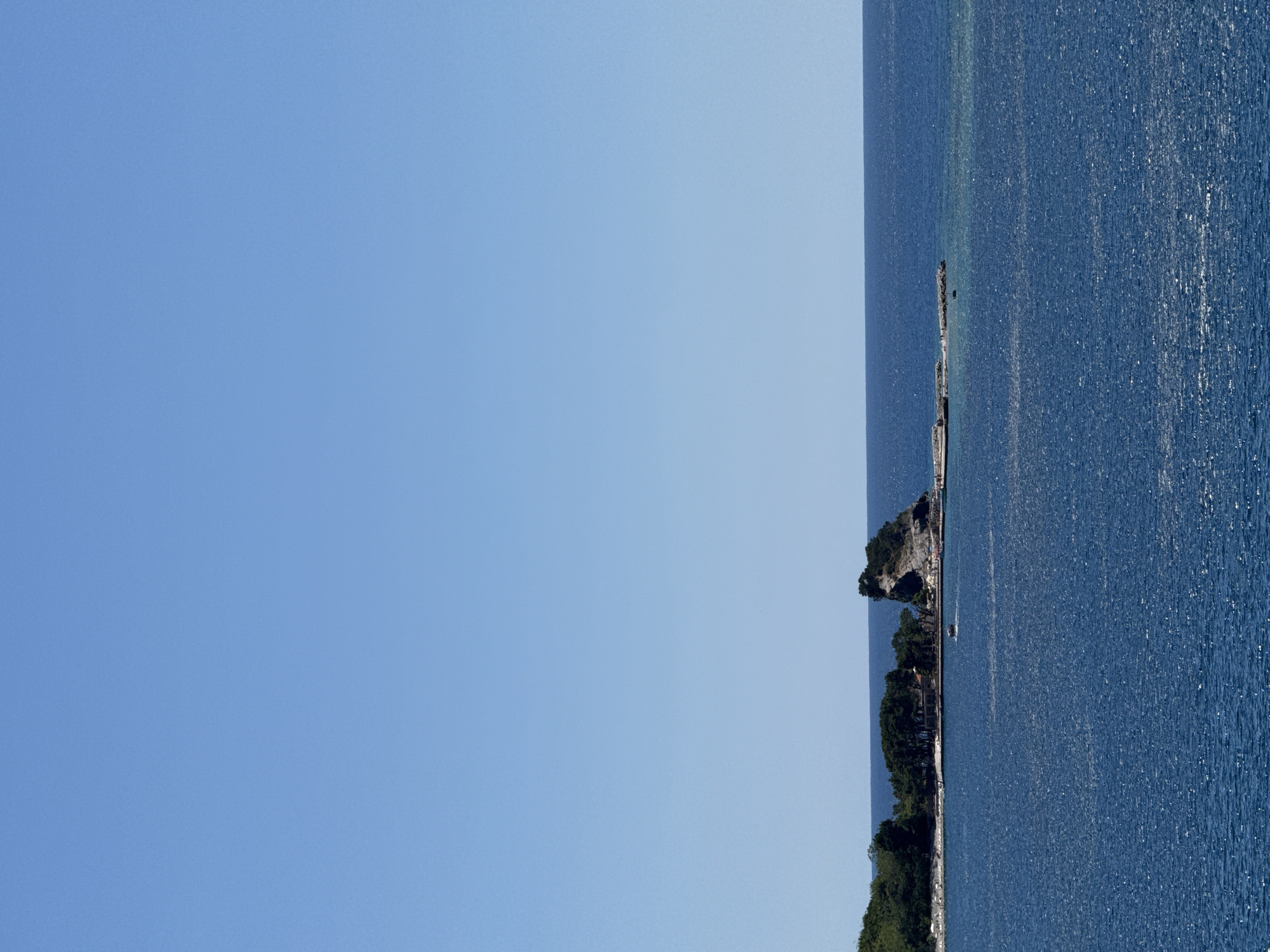 The image depicts a scenic view of the coastline near Budva, Montenegro, under a clear, bright blue sky. The foreground is dominated by the deep blue, shimmering waters of the Adriatic Sea, with sunlight creating a sparkling effect on its surface. In the middle ground, a small, rocky island rises from the water, topped with rugged formations and some vegetation. Adjacent to this island, a narrow, paved walkway or pier extends into the sea, with several people visible walking along it. Further to the left, a larger landmass is covered with lush green trees and some structures, suggesting a coastal town or resort area. A small boat is seen moving through the water between the mainland and the island. The horizon line is sharp, separating the sea from the vast, cloudless sky. The overall impression is one of a tranquil and sunny day by the sea in a picturesque coastal setting.