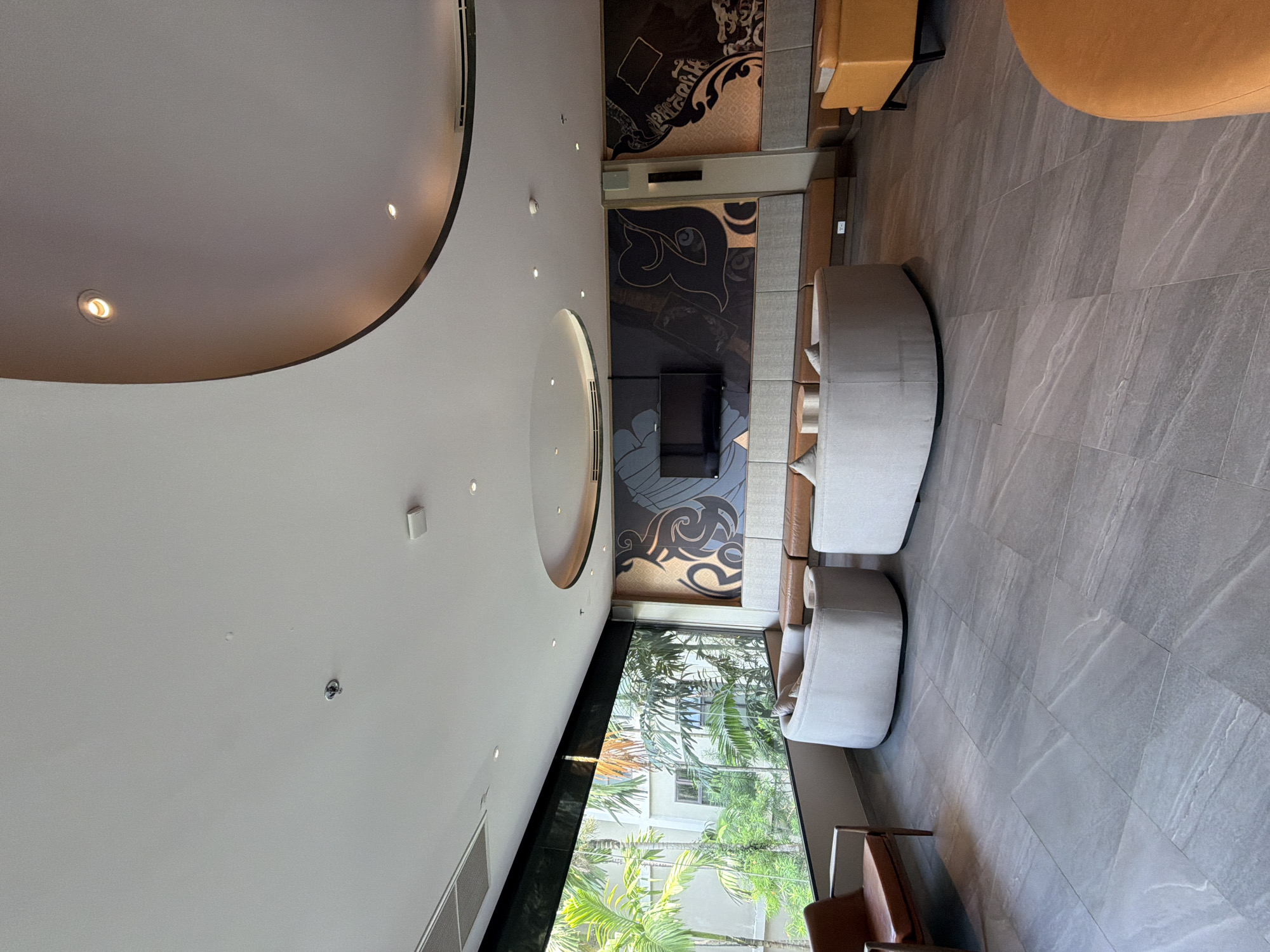 This image depicts a spacious, modern lounge area with large windows offering a view of lush tropical greenery and a building exterior. The room features a gray tiled floor. In the foreground, two cream-colored, curved sofas are arranged. To the left, a brown leather armchair is visible. The back wall is adorned with a large, stylized mural in dark colors and gold accents, with a black television mounted centrally. A continuous built-in sofa with brown and gray upholstered sections runs along the back wall. To the right, a bright orange armchair is positioned. The ceiling is white with recessed lighting and a curved architectural element. This appears to be a hotel lobby or common area in Bang Muang, Thailand, given the visual cues of tropical foliage and architectural style often found in resorts. There are no people visible in the scene.