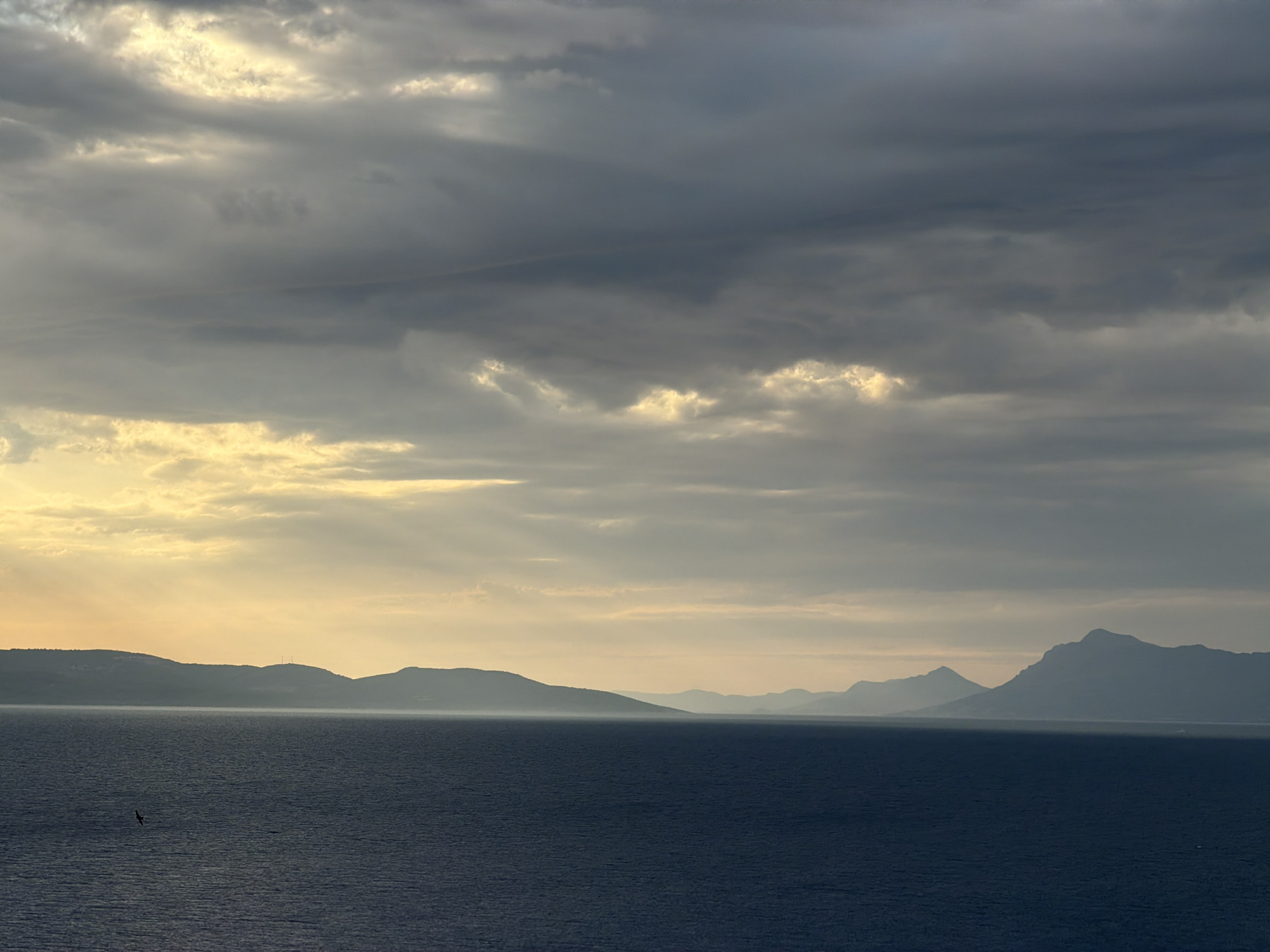 The image depicts a serene seascape at dusk, with dramatic, cloudy skies illuminated by the golden hues of the setting sun. Silhouetted mountains form a distant coastline, receding into a hazy horizon. The dark, calm water in the foreground is dotted with small waves, and a lone bird is captured in flight, adding a sense of scale and movement to the vast expanse. The scene evokes a tranquil and contemplative atmosphere, characteristic of the natural beauty found along the Dalmatian coast. The location is Općina Podgora, Croatia, a coastal municipality in the Split-Dalmatia County.