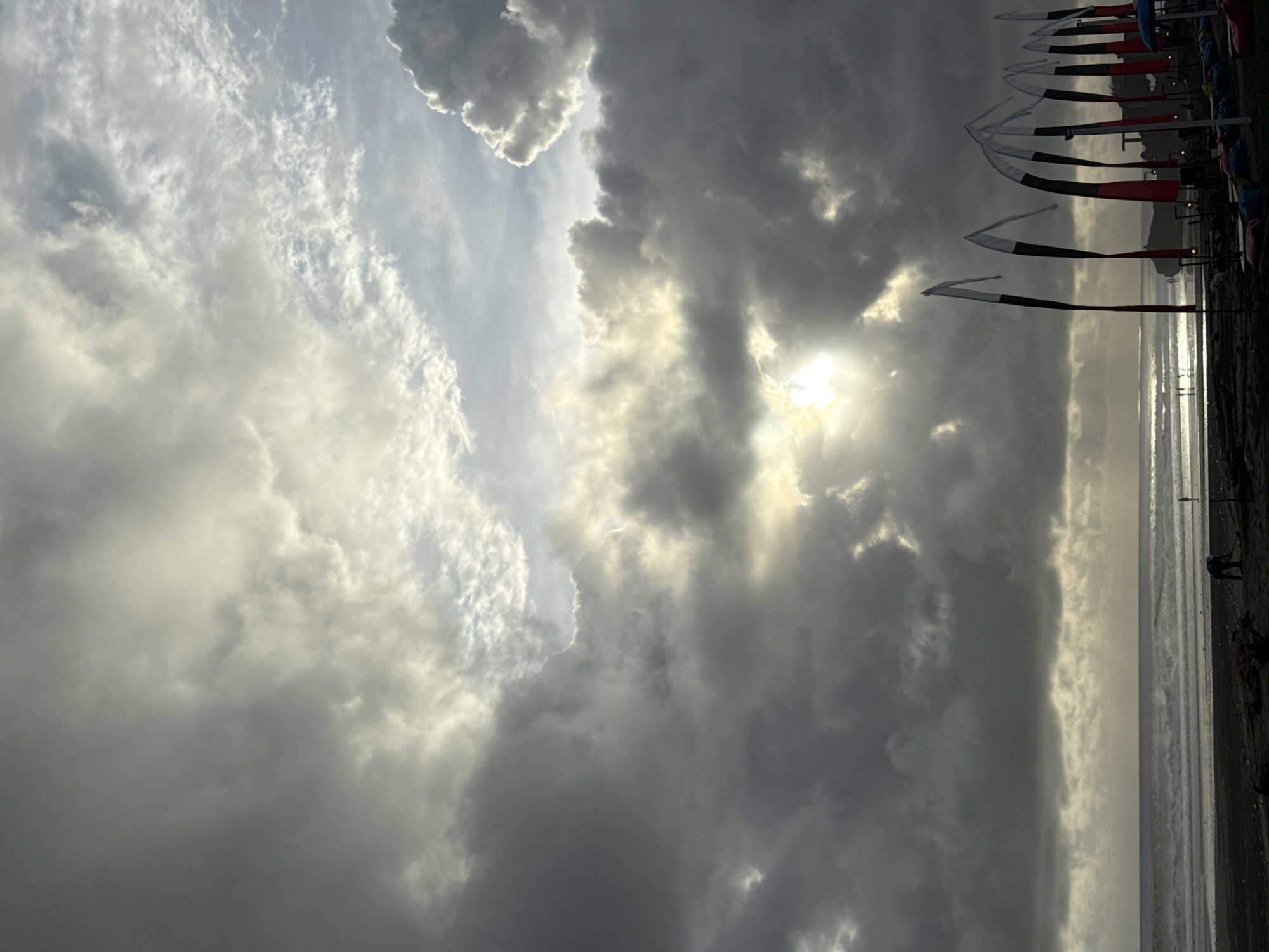 A partially obscured sun shines over the sea and a beach. The sky features dark gray storm clouds, with a patch of lighter, higher altitude clouds to the left. Flags are visible on the right, near the beach. The location is unknown.
