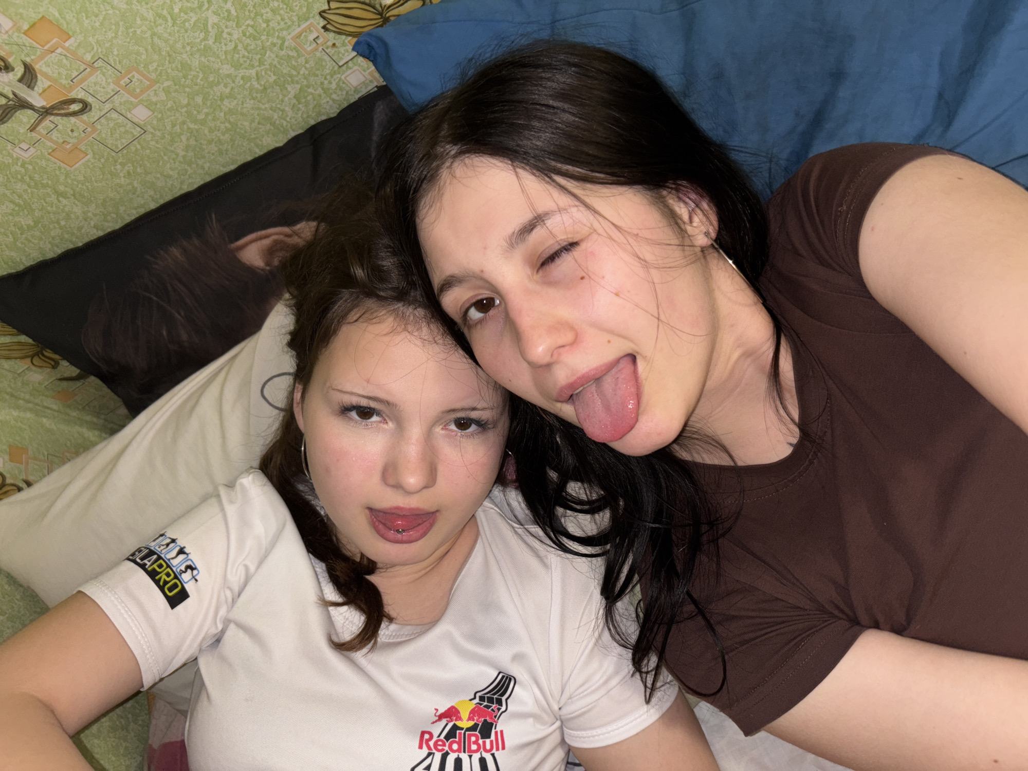 Two young women are lying down on what appears to be a bed. The woman on the left is wearing a white t-shirt with a "Red Bull" logo and the word "LAPRO" printed on the sleeve; she has light skin, light brown hair and is sticking her tongue out. The woman on the right is wearing a brown t-shirt, has dark hair, is winking, and is also sticking her tongue out. A blue pillow is behind the woman on the right. A green bedspread with a geometric pattern is visible on the left. A black pillow is behind the woman on the left. The location is unknown and Фоки, Russia cannot be determined.