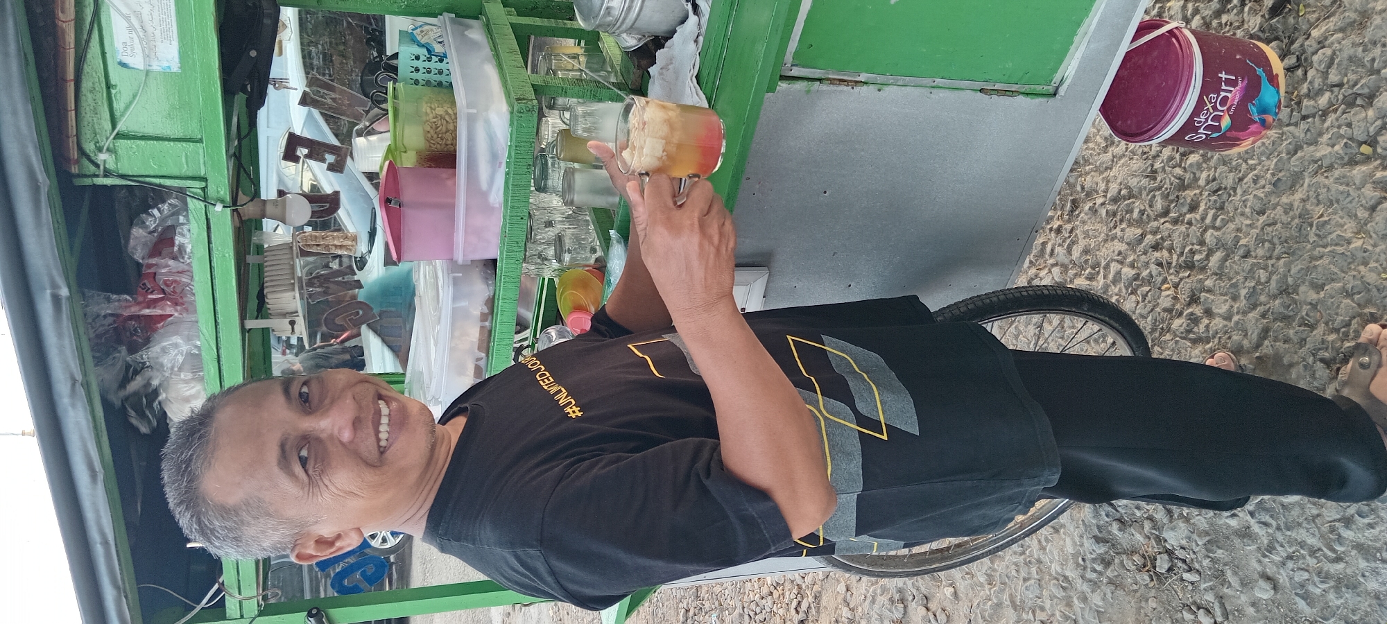 A middle-aged Indonesian man with short, graying hair and a weathered face smiles broadly at the camera, holding a glass of iced beverage. He wears a black t-shirt with a yellow and gray geometric pattern and the hashtag #UNLIMITEDJOURNEY. The man stands beside a mobile food cart, painted green, which is stocked with various items in plastic containers. Behind him, a bicycle wheel is visible. On the ground, near the cart, is a purple paint bucket labeled "Dexa Smart Emulsion Paint." The ground is covered in rough gravel and small stones. The background suggests an outdoor setting, possibly a street or market in Kalipucang, Indonesia, with other carts and vehicles blurred in the distance.