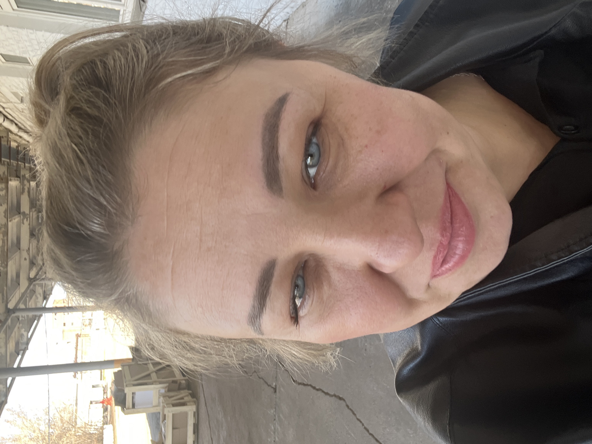 A close-up, slightly tilted selfie shows a fair-skinned woman with blonde hair and blue eyes. She has visible freckles and her lips are pink. She is wearing a black leather jacket. The background shows a gray concrete surface, a metal structure and wooden crates. The location is in Chaikovsky, Russia.