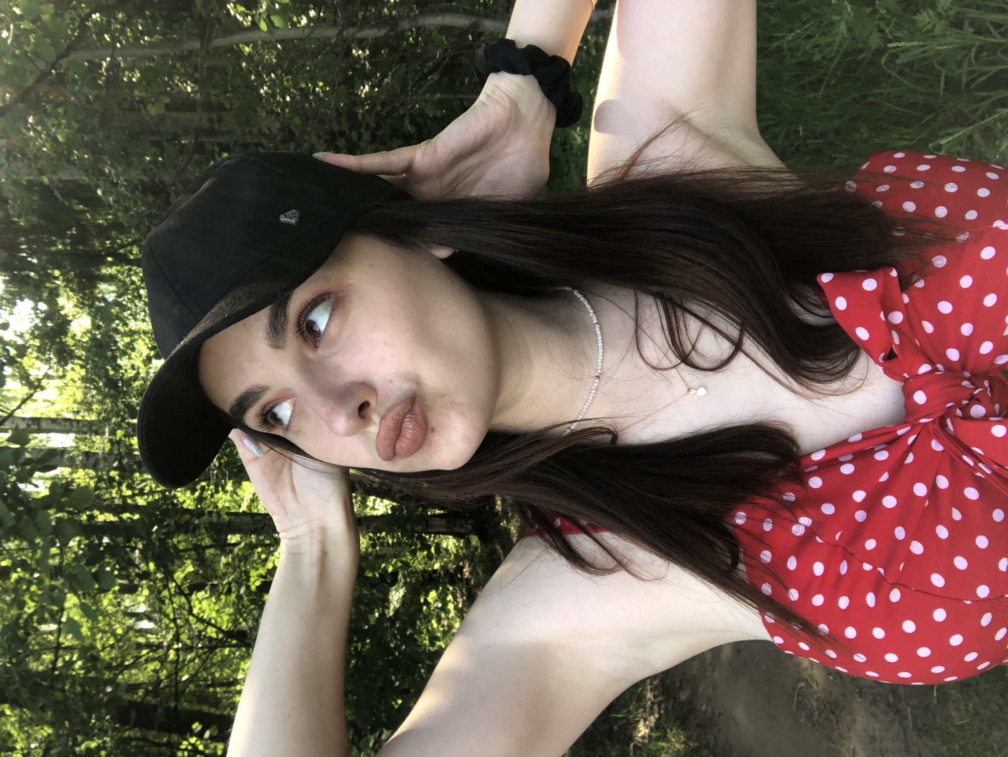 A woman is outdoors, likely in a park or wooded area. She wears a black baseball cap, a red dress with white polka dots, and a delicate beaded necklace. Her dark hair is loose around her shoulders. She's posed with her hands near her head, wearing a scrunchie on her right wrist. She appears to be in Noviy, Russia, based on analysis of visual details and landmarks. The environment includes trees and foliage, suggesting a natural setting.