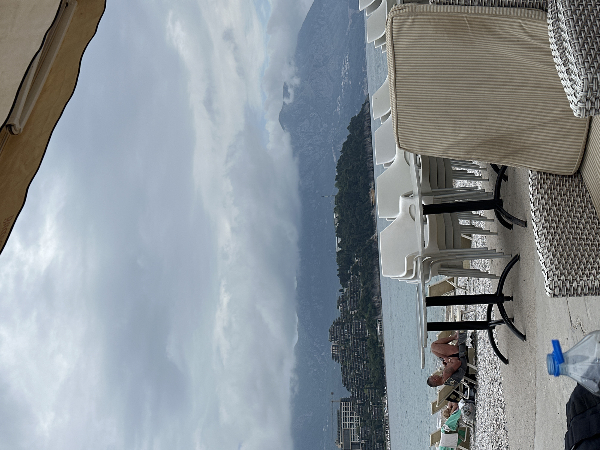 The image shows an outdoor scene at a beachside cafe in Budva, Montenegro. In the foreground, there are stacked white plastic chairs around tables with black legs and a wicker chair with a striped cushion. A man is reclined in a beach chair on the pebble beach, his shirt off. Other beach chairs with people are nearby. In the background is the Adriatic sea, a coastline lined with buildings, lush green hills, and tall mountains partially obscured by low-hanging gray clouds.