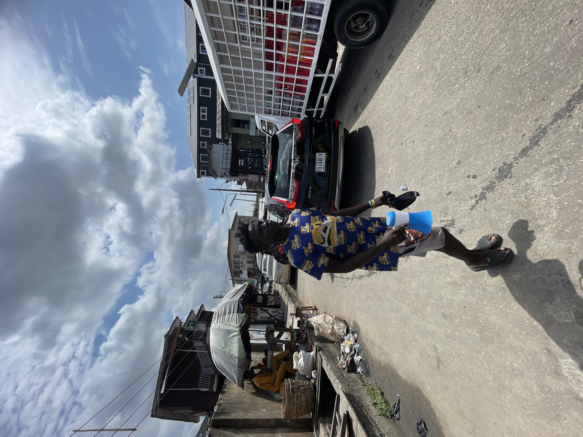 This image depicts a bustling street scene in Lagos, Nigeria, under a bright, partially cloudy sky. The primary subject is a dark-skinned man walking forward on the right side of the frame. He wears a blue short-sleeved shirt patterned with yellow motifs that include the text "Burger's" and possibly a stylized "Y" or animal. He is also wearing light-colored shorts and black slides. In his left hand, he holds a blue and white megaphone along with a black item, possibly a small bag or speaker. He carries a white plastic bottle in his right hand.
The strong sunlight casts long, distinct shadows of the man and other elements on the concrete pavement, suggesting it is midday or early afternoon. On the street behind him, a black Honda CR-V is visible, bearing the license plate "FJ 448JV." Behind the car is a large white truck with a caged back, filled with numerous red crates, likely used for transporting goods or beverages. Multi-story buildings line the street in the background.
On the left side of the pavement, a street vendor's stall with a white umbrella is set up. A person in yellow or orange attire is seated behind the stall, next to a woven basket. Piles of trash and debris are visible along the edge of the pavement, near what appears to be a gutter or drainage system. The sky above is a mix of blue patches and scattered white clouds. The scene conveys a sense of daily life and commerce in an urban setting.