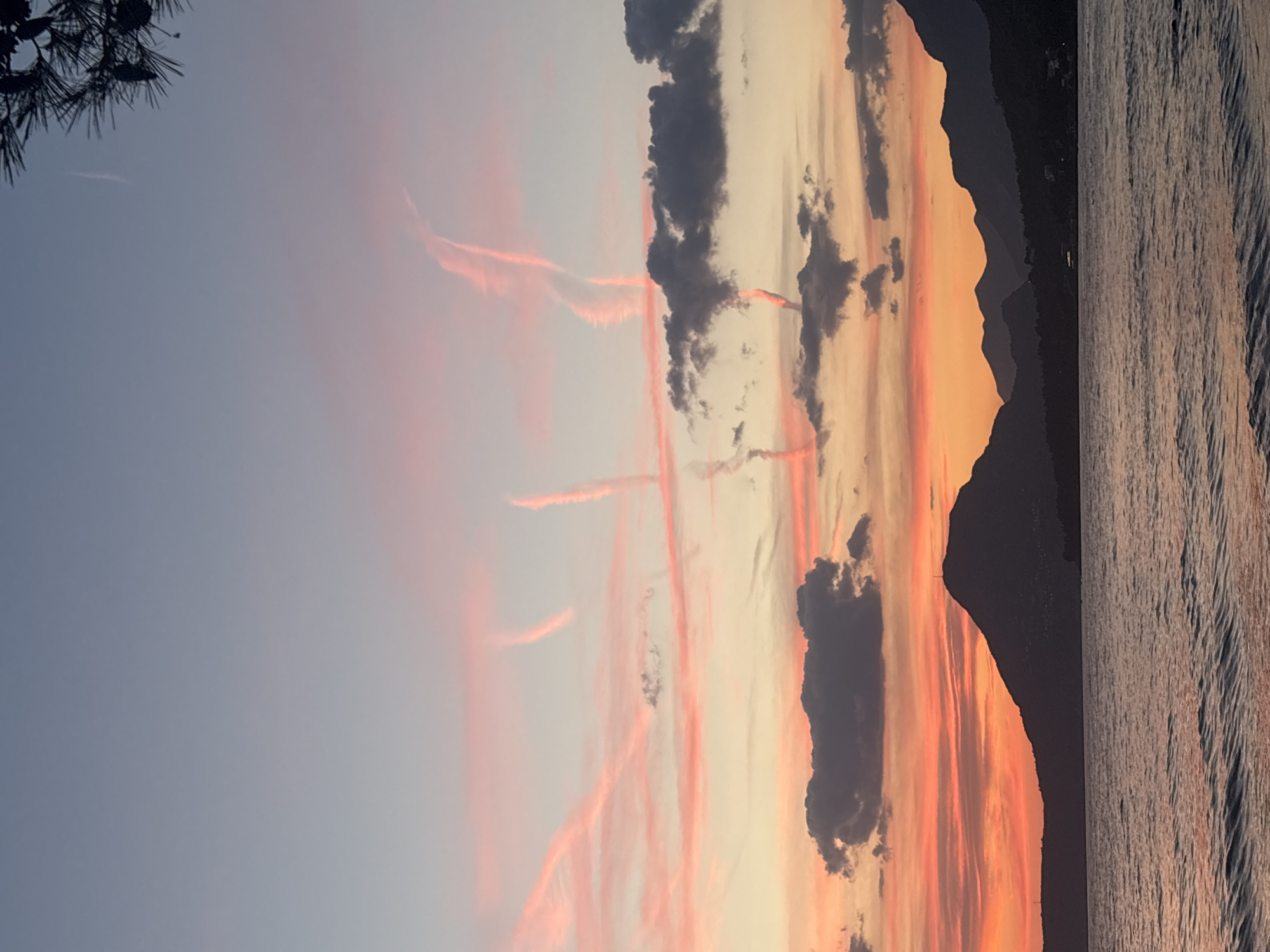 The image captures a sunset scene in Bar, Montenegro. The right edge shows water rippling, reflecting the sunset colors. Adjacent to the water is a silhouetted mountain range. The sky above is a gradient, transitioning from pale blue to orange and pink. Contrails are visible against the blue backdrop. There are dark clouds scattered throughout the center of the frame. A portion of a tree with pine needles is visible in the top left corner.