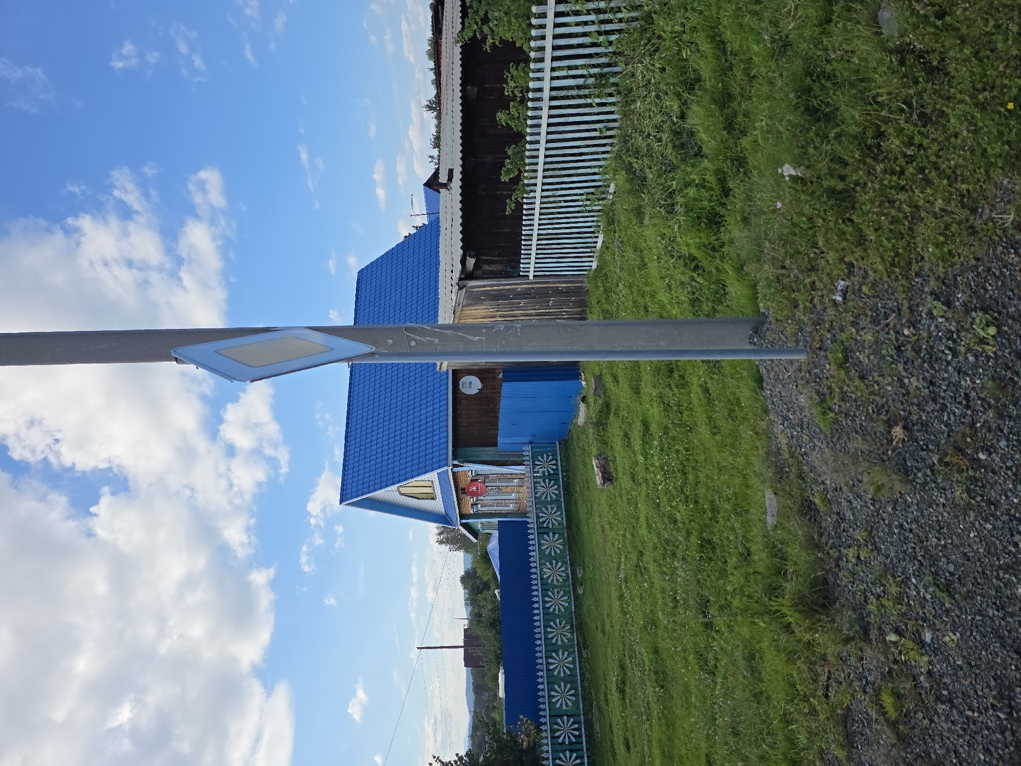 This is a wide, eye-level, outdoor, daylight shot of a rural Russian village in Yuzhny Gornyak. The main subject is a wooden house with a bright blue, pitched roof. The house has a gable end with a window and wooden siding. In front of the house is a blue fence with a decorative white pattern. To the right of the house is a wooden structure with a corrugated metal roof, enclosed by a white picket fence. A utility pole with a diamond-shaped road sign is in the foreground, to the right of the center, obscuring part of the house and structures. The ground is covered in green grass and gravel. The sky is blue with scattered white clouds.