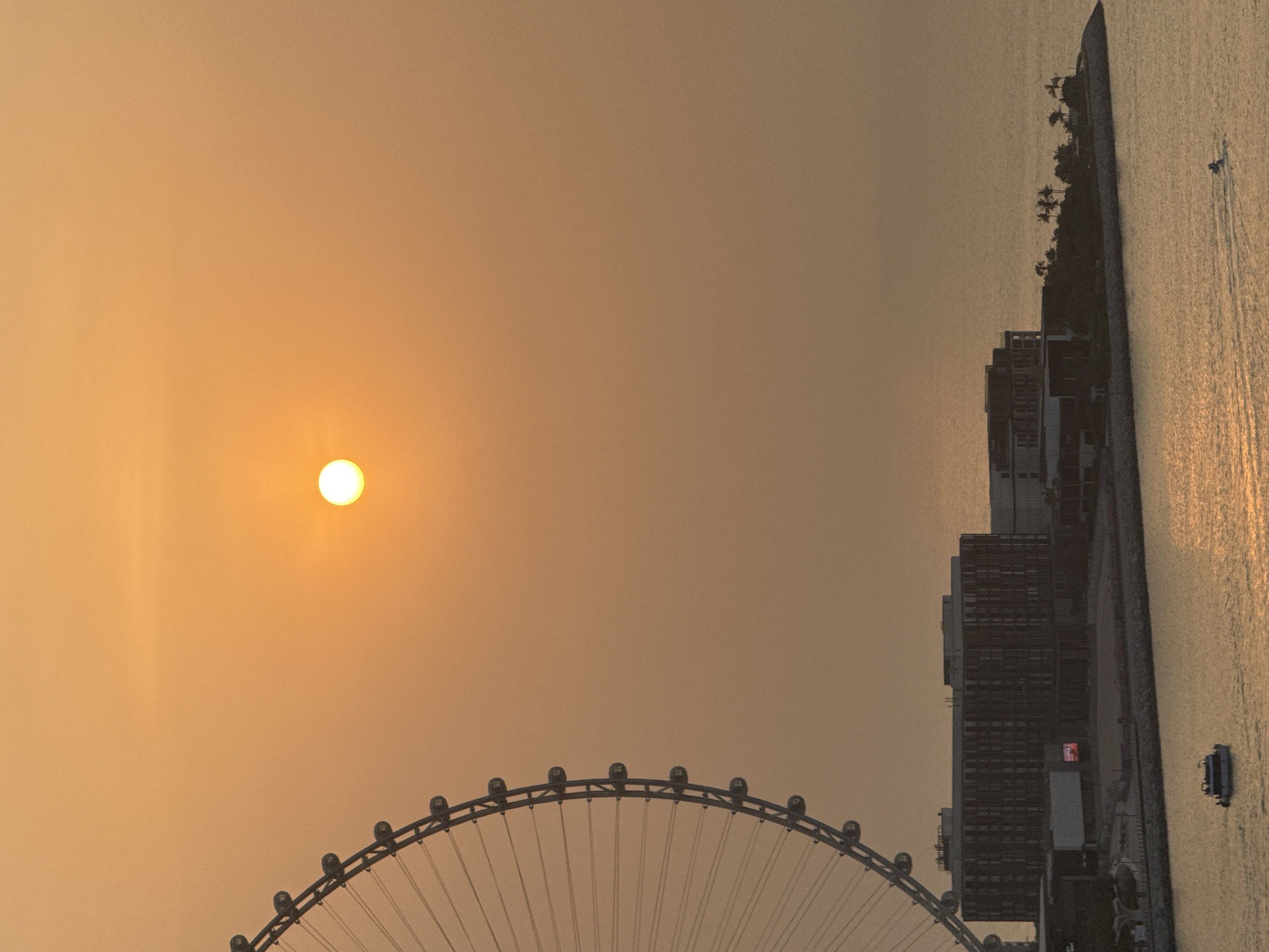 The image shows a sunset over the Arabian Gulf in Dubai, United Arab Emirates. The sun is a bright, glowing orb in the hazy orange sky. On the left, a large Ferris wheel, likely Ain Dubai, dominates the frame. In the middle ground, a modern hotel complex with dark facades and multiple floors is visible along the coastline, with a large screen displaying a red illuminated advertisement. The sea is calm, reflecting the warm hues of the sunset. Two small boats are visible on the water: one larger white boat is closer to the shore, and a smaller jet ski is further out, leaving a wake. The coastline is lined with palm trees, indicating a tropical or subtropical environment. The overall scene evokes a sense of tranquility and the end of a day in a vibrant city.