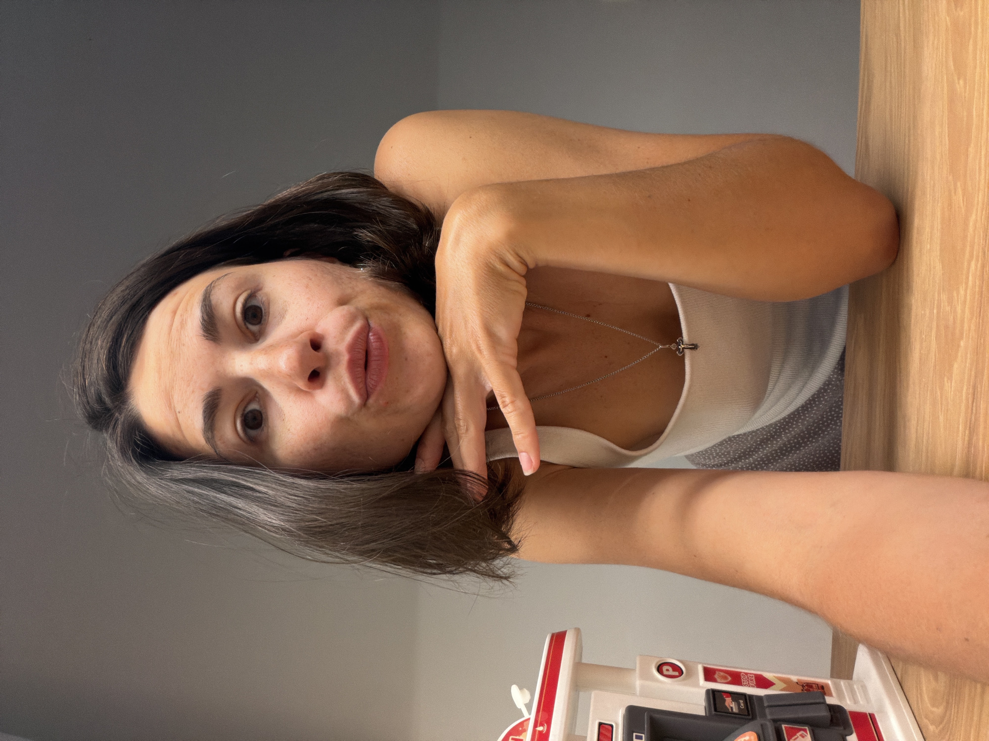 A woman with brown hair and fair skin is shown in a selfie. She is wearing a white tank top and a silver necklace with a cross pendant. Her left hand is placed near her face, which is tilted slightly towards the camera. To the right of the frame, a wooden surface is visible. In the lower portion of the image, a toy gas station with red and white coloring is partially visible. The background is a neutral gray wall. The location is not identifiable as Budva, Montenegro.