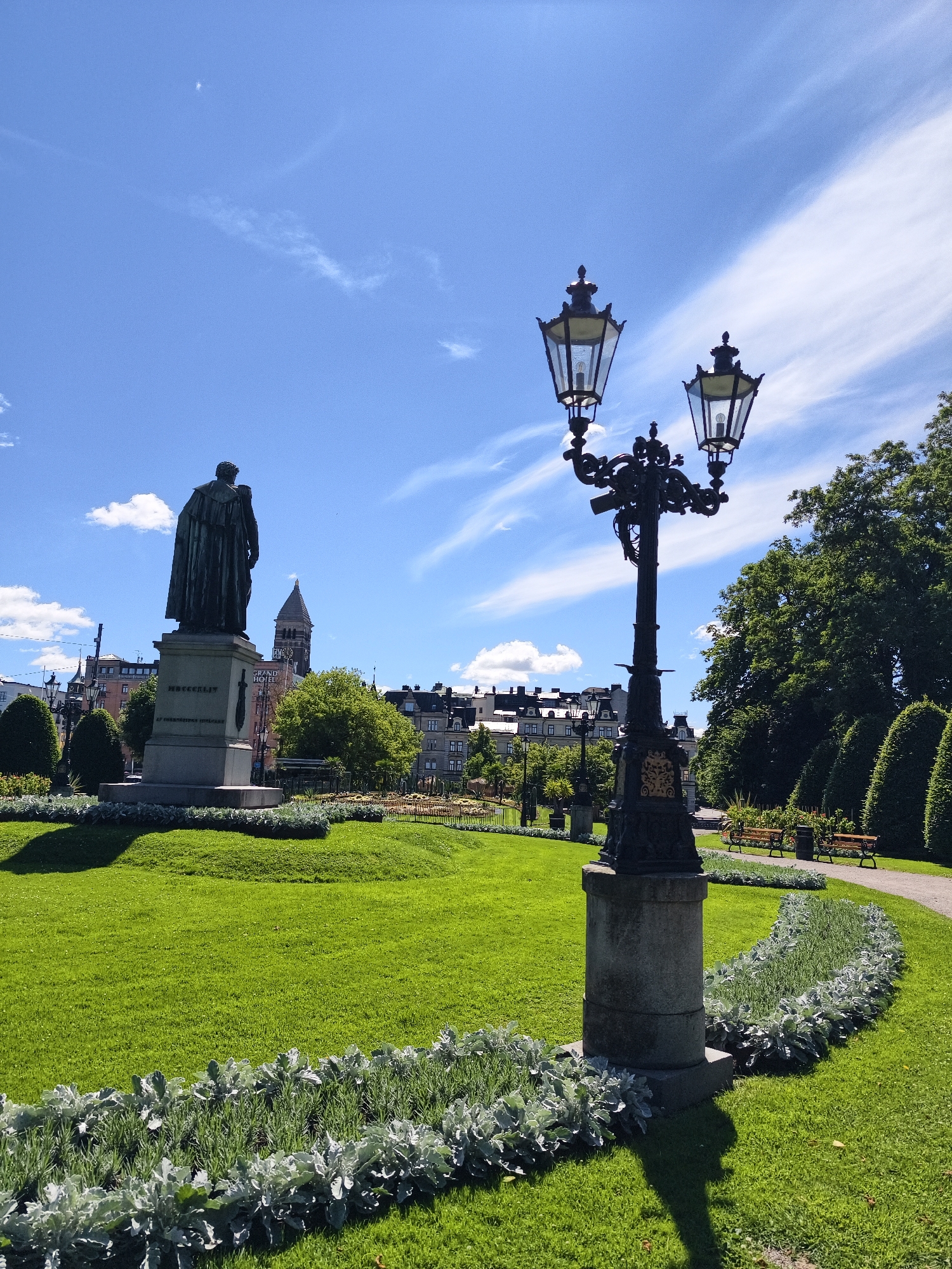 The image shows a statue of a man in a long coat, facing away from the camera, in a city park. The statue is on a stone pedestal with inscriptions. In the foreground, there is a lamppost with two lanterns. The park has manicured green lawns, flower beds, and trees. In the background, there are buildings and a church tower. The sky is blue with some white clouds. The scene appears to be in Norrköping, Sweden, as indicated by the statue's inscription which reads "L.M.L." followed by "MDCCXLVII" (1747), referring to Lars Magnus Ljungberg, a prominent figure in Norrköping's industrial history and a city council member, who lived from 1778 to 1847. The statue is located in Gamla Rådstugugatan 6, Norrköping, Sweden, which is near the city center and the industrial heritage area.