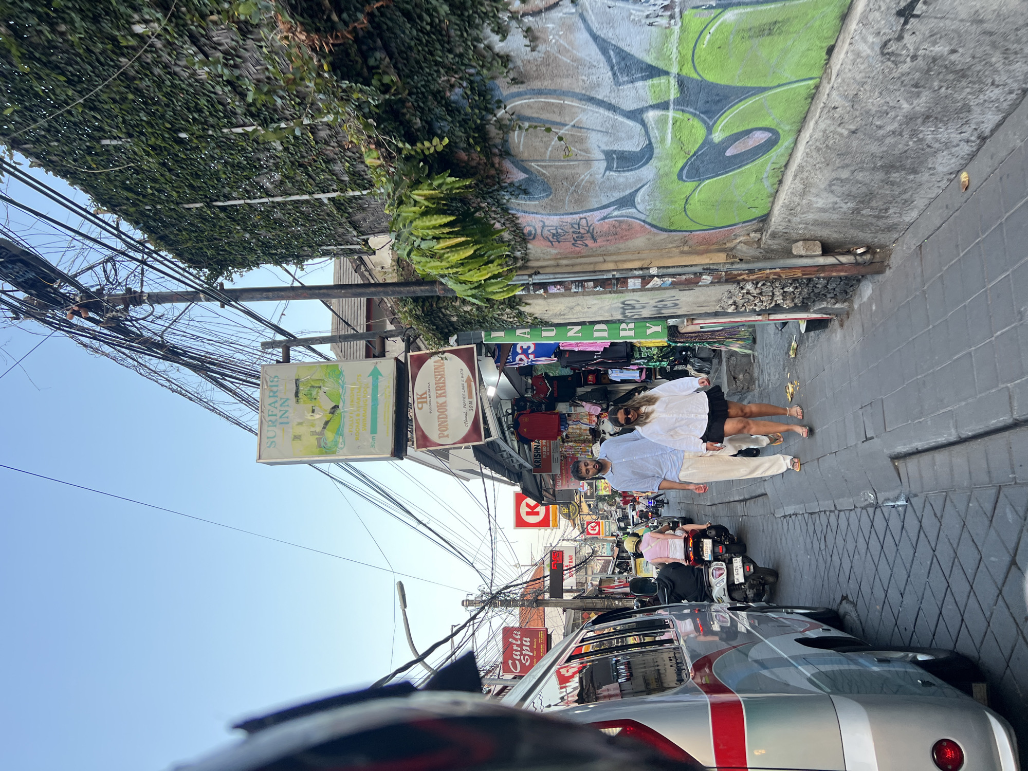 This image captures a bustling street scene in Kuta, Indonesia, likely a commercial area with a mix of shops, accommodations, and vehicle traffic. In the foreground, the side of a silver and red bus is visible on the left. A young couple walks towards the viewer on the right side of the narrow street. The man, wearing a light blue button-up shirt and light-colored pants, walks slightly ahead of the woman, who is in a white long-sleeved shirt and a black skirt.
The street is paved with interlocking gray bricks. Several motorcycles are present, with one occupied by a person wearing a pink shirt and helmet, and another with a person in a black shirt. Overhead, a dense network of electrical wires crisscrosses the sky. Signs for "SURFARI INN" and "PONDOK KRISHNA" are visible, indicating nearby businesses. A building on the right is covered in dense green ivy. Graffiti art, predominantly in green and white, adorns the concrete wall on the right side of the street. Shops with clothing racks and various items are set up along the sidewalk on the right. The overall environment suggests a vibrant and busy tourist destination.