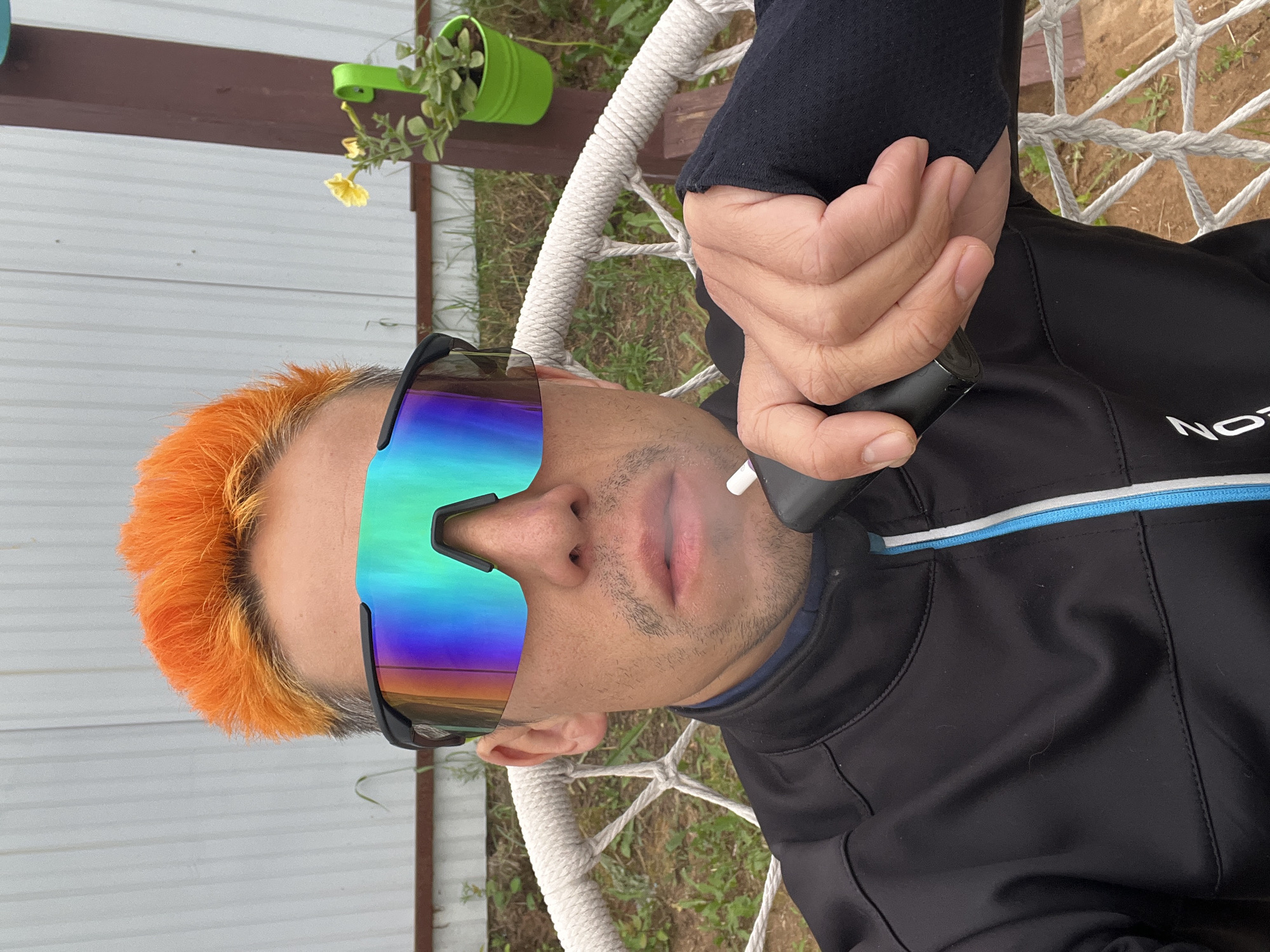 A selfie taken outdoors in Chaikovsky, Russia shows a man with bright orange hair sitting in a white rope chair. He is wearing large, iridescent, wrap-around sunglasses and a black jacket with blue and white trim. The man is holding a dark electronic smoking device to his mouth, from which a plume of vapor is emanating. In the background, a light-colored, corrugated metal wall is visible, along with some greenery and a small plant in a bright green pot.