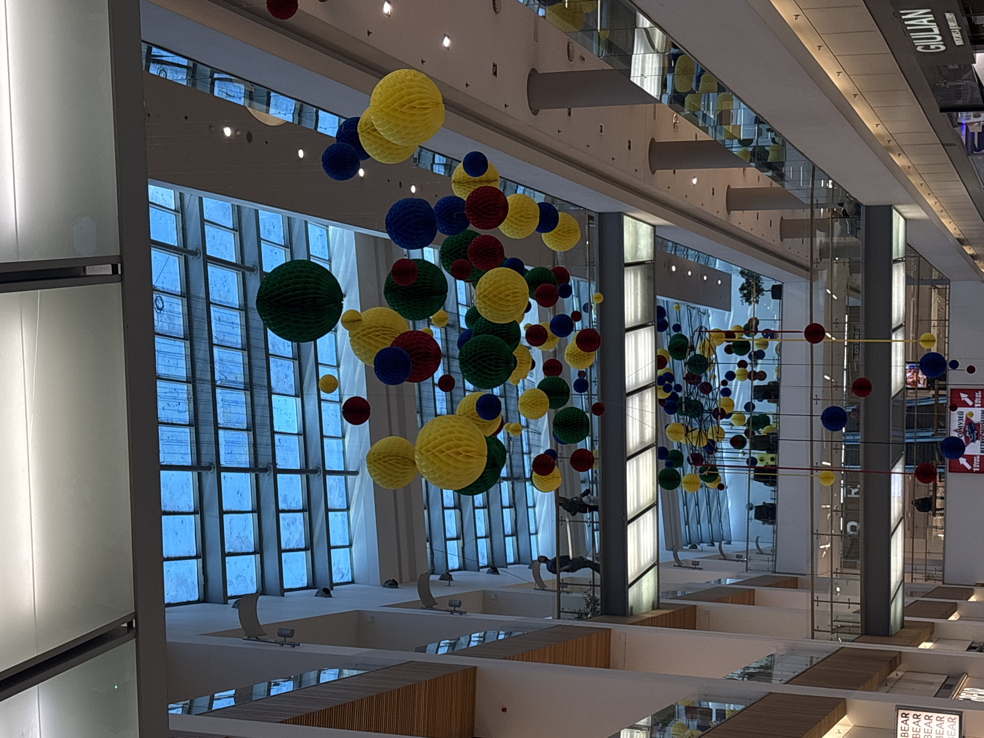 The image shows an interior view of the Grand Mall Varna in Varna, Bulgaria. The scene is a multi-story atrium with large windows on one side. Decorative hanging orbs, in red, blue, green, and yellow, are suspended from the ceiling. The mall's design includes visible levels and walkways with wooden accents. There is no readily visible people in the image. The environment appears to be well-lit and the overall atmosphere is modern and commercial.
