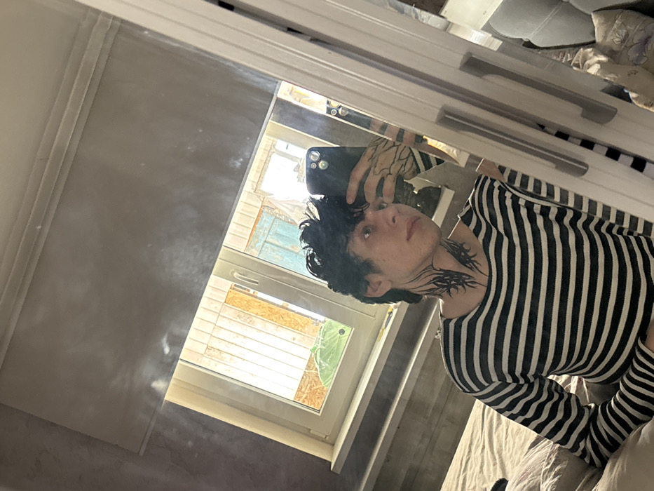 This image displays a young male taking a mirror selfie inside a room. The specific city of Tatishchevo, Russia, cannot be visually confirmed from the image content.
The subject has dark, curly hair and is wearing a long-sleeved, horizontal striped shirt (dark and light). He exhibits distinct black tribal-style tattoos on both sides of his neck and patterned tattoos, resembling branches, on his visible right forearm. He holds a dark-colored smartphone, likely an iPhone due to the visible camera array, with his left hand, partially obscuring his face. His gaze is directed slightly away from the phone's lens, towards his reflection.
He is positioned on a bed with light-colored bedding. The mirror used for the selfie features a light-colored frame and two silver horizontal handles on the right side, suggesting it is part of a piece of furniture like a wardrobe or closet door. Behind the subject, a window with a white frame is visible, partially covered by a grey roller blind. Through the window, an exterior view includes parts of a building or structure with exposed wooden elements and a segment of a light blue painted surface. A large, bright green moth, possibly a Luna Moth, is visible on or near the windowsill. The interior wall to the left of the window exhibits a textured, grey finish. The room is illuminated by natural light entering through the window.