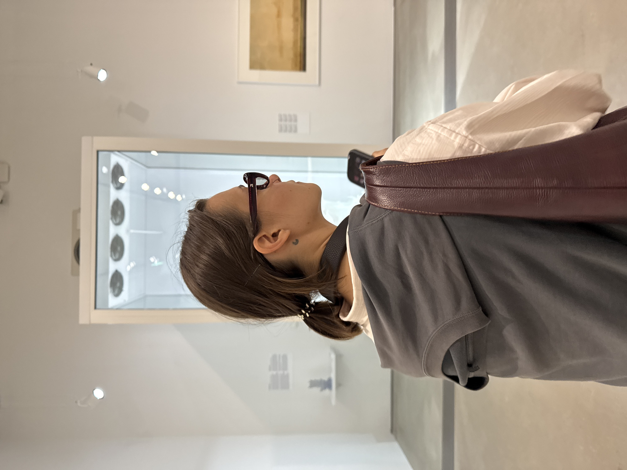 A medium shot captures a woman from behind, facing right in profile, in a museum. She has dark hair pulled back into a ponytail and wears large, dark-rimmed glasses. A small crescent moon tattoo is visible on her neck. She is dressed in a white collared shirt under a dark grey hooded vest. A burgundy leather purse is slung over her right shoulder. Her right hand is holding a smartphone. The background suggests an art exhibition with white walls, track lighting, a glass display case, and framed artwork. The floor is concrete.