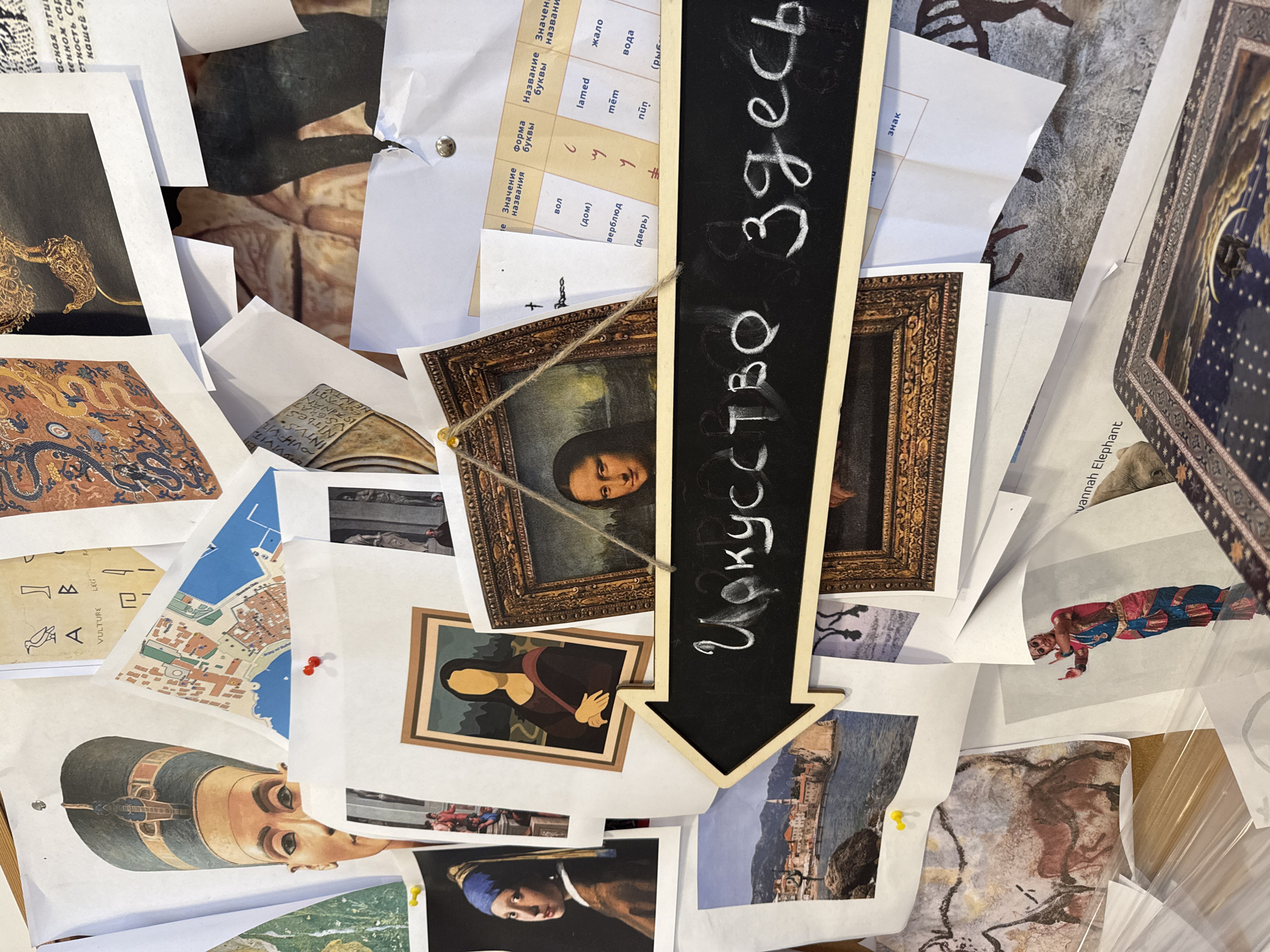 A chaotic assortment of art-related images is spread out on a surface, possibly a table or board. The images include reproductions of famous artworks such as Leonardo da Vinci's Mona Lisa, the Girl with a Pearl Earring, ancient Egyptian art, cave paintings, and various cityscapes. A black chalkboard arrow with the word "Искусство здесь" written on it is positioned amongst the images, pointing downwards. The phrase translates to "Art is here." A map of a city is visible, pinned with two red tacks, along with a print of a cityscape showing the location in the city of Budva, Montenegro. The composition suggests a collage or mood board related to art or an art project.