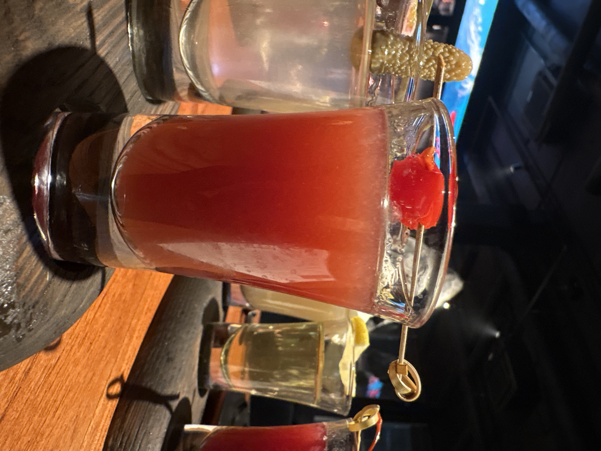 The image shows a wooden tray holding several small glasses of what appear to be different alcoholic beverages. The most prominent glass contains a red drink garnished with a red cherry or tomato on a skewer. Other glasses contain a clear liquid, one with a pickle garnish, and another with a lemon wedge. The setting appears to be a dimly lit bar or restaurant, possibly in Anapa, Russia, judging by the request. No people are visible in the frame. The focus is on the drinks and the presentation on the wooden tray.