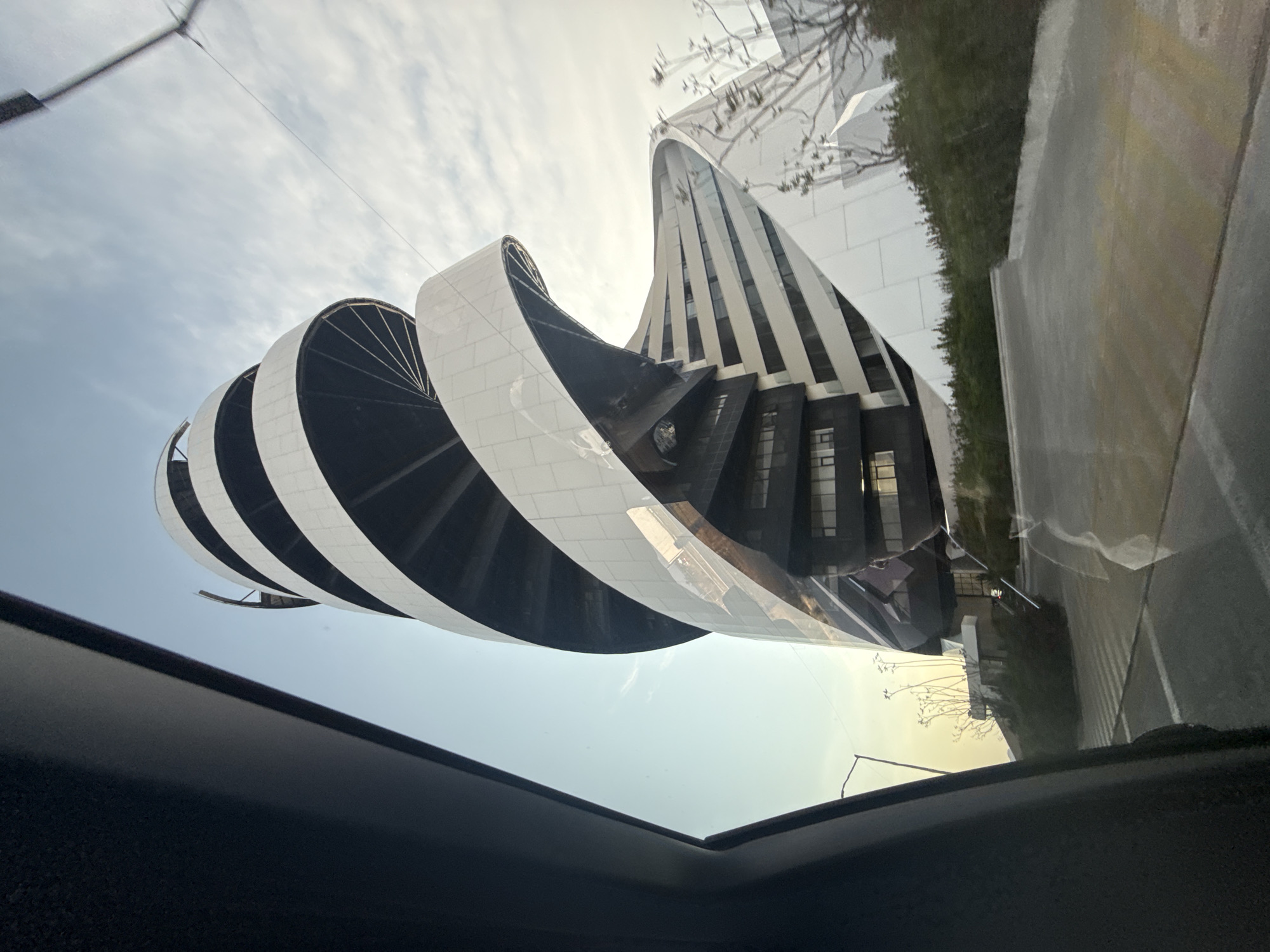 The image shows a view from inside a vehicle, looking up at the Solarium Tower located in San Bernardino Tlaxcalancingo, Mexico. The tower features a cylindrical design with alternating white and black horizontal bands. Adjacent to the Solarium Tower is another building that has a modern architectural style with white vertical stripes. A road and greenery are visible to the side. The sky is partially visible, with some cloud cover.