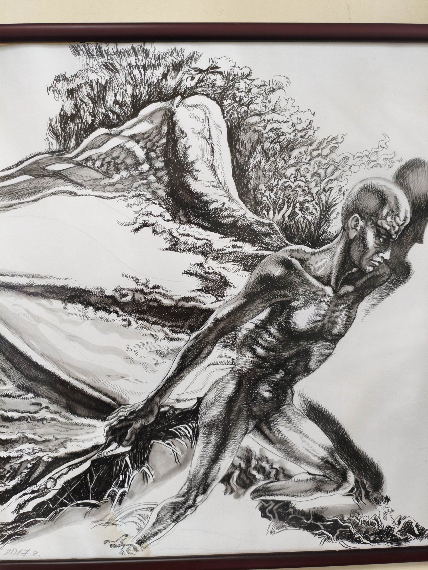 The image is a framed ink drawing of a muscular, bald man with wings, possibly an angel or fallen angel, depicted in a dynamic, struggling pose. He is reaching down, his hands gripping what appears to be fabric or a natural element, perhaps earth or water. The background is abstract and suggestive, with swirling lines and textures that could represent wind, smoke, or turbulent nature. The drawing style is detailed and dramatic, using strong contrasts of light and shadow. The lower left corner of the artwork shows a signature and date: "2017 2.". The artwork is framed with a dark reddish-brown frame. There is no discernible location in the city of Burgas, Bulgaria within the artwork itself.