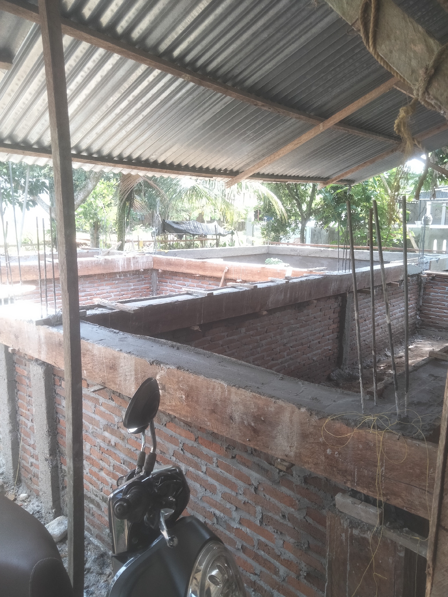 The image shows a construction site in Kruengmanyang, Indonesia. The foundation of a building is under construction, with red brick walls and concrete beams. Rebar rods protrude vertically from the concrete. The construction is covered by a corrugated metal roof supported by wooden beams. A black motorcycle is parked in the foreground. Trees are visible in the background outside the construction area.