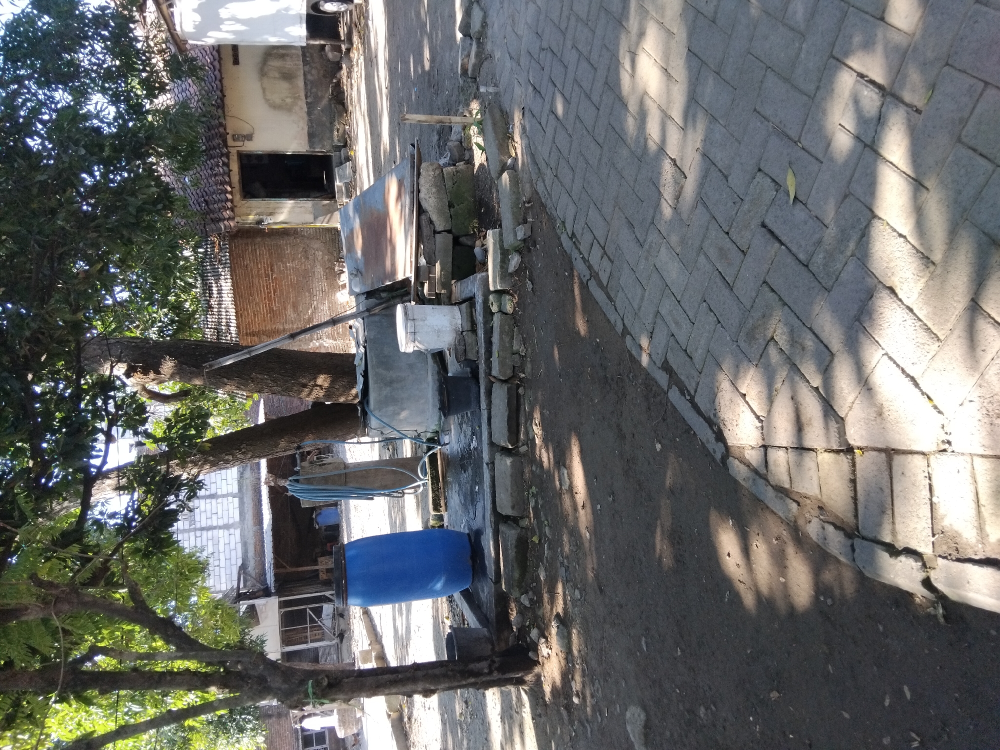 In the city of Kepuh Kiriman, Indonesia, an outdoor scene depicts a well or water collection area with several objects. A large blue plastic barrel stands prominently on the left, next to a smaller, dark-colored bucket. To the right of the barrel is a concrete structure with a metal lid, appearing to be a well or cistern. A blue hose is coiled and draped over a wooden pole near this structure. Two black buckets are positioned near the well. Behind these objects, a building with a tiled roof and brick walls is visible. In the foreground, a paved walkway made of interlocking bricks runs diagonally across the image, with dirt and debris in the surrounding ground. Trees with leafy canopies cast shadows on the scene, suggesting a daytime setting. No people are visible in the frame.