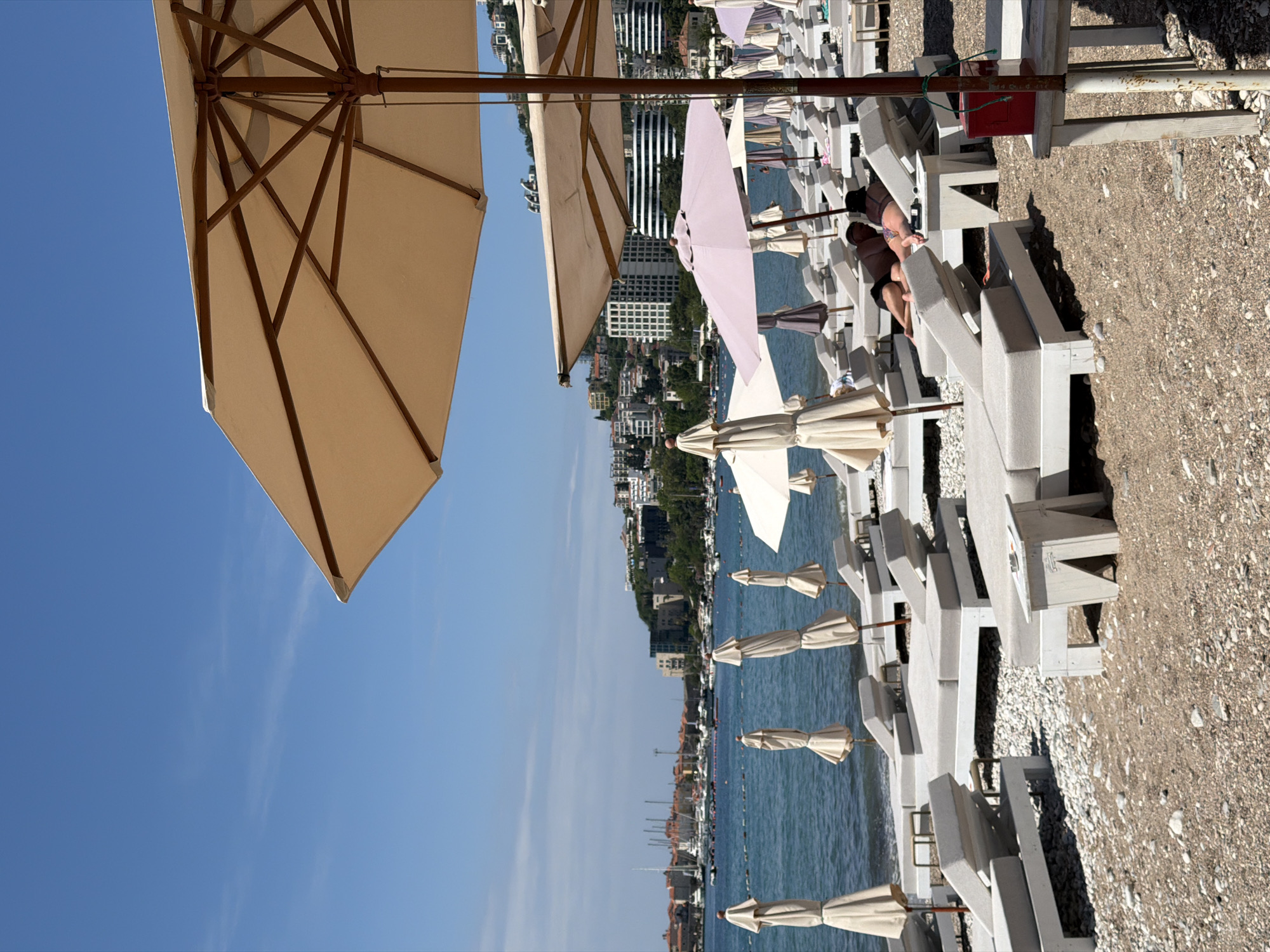 This media file captures a sunny day at a beach resort in Budva, Montenegro, with a clear blue sky overhead. The image is taken from a low angle, looking across a beach towards the sea and the coastal town.
In the foreground, numerous light-colored, possibly concrete or stone, sun loungers are arranged in rows on a pebbly beach. Accompanying these are many beach umbrellas, mostly of a beige or cream canvas material; some are open, providing shade, while others are neatly closed and tied. One distinctive light pink or purple open umbrella is visible among the beige ones. A person can be seen lying relaxed on their back on a sun lounger, wearing dark clothing, with a phone resting beside them. Another pair of legs is visible further down a row of loungers. A red box, possibly for electrical access, is attached to an umbrella pole.
Beyond the stretch of beach, the calm, blue-green sea is dotted with various boats and yachts. The background features the coastal town of Budva, with a mix of multi-story buildings, some modern and some older, nestled against green hills and vegetation. The scene suggests a busy but relaxed leisure activity, typical of a summer day at a Mediterranean destination.
The time of day appears to be mid-morning or early afternoon, indicated by the bright sun and clear lighting. There is no discernible text visible in the image.