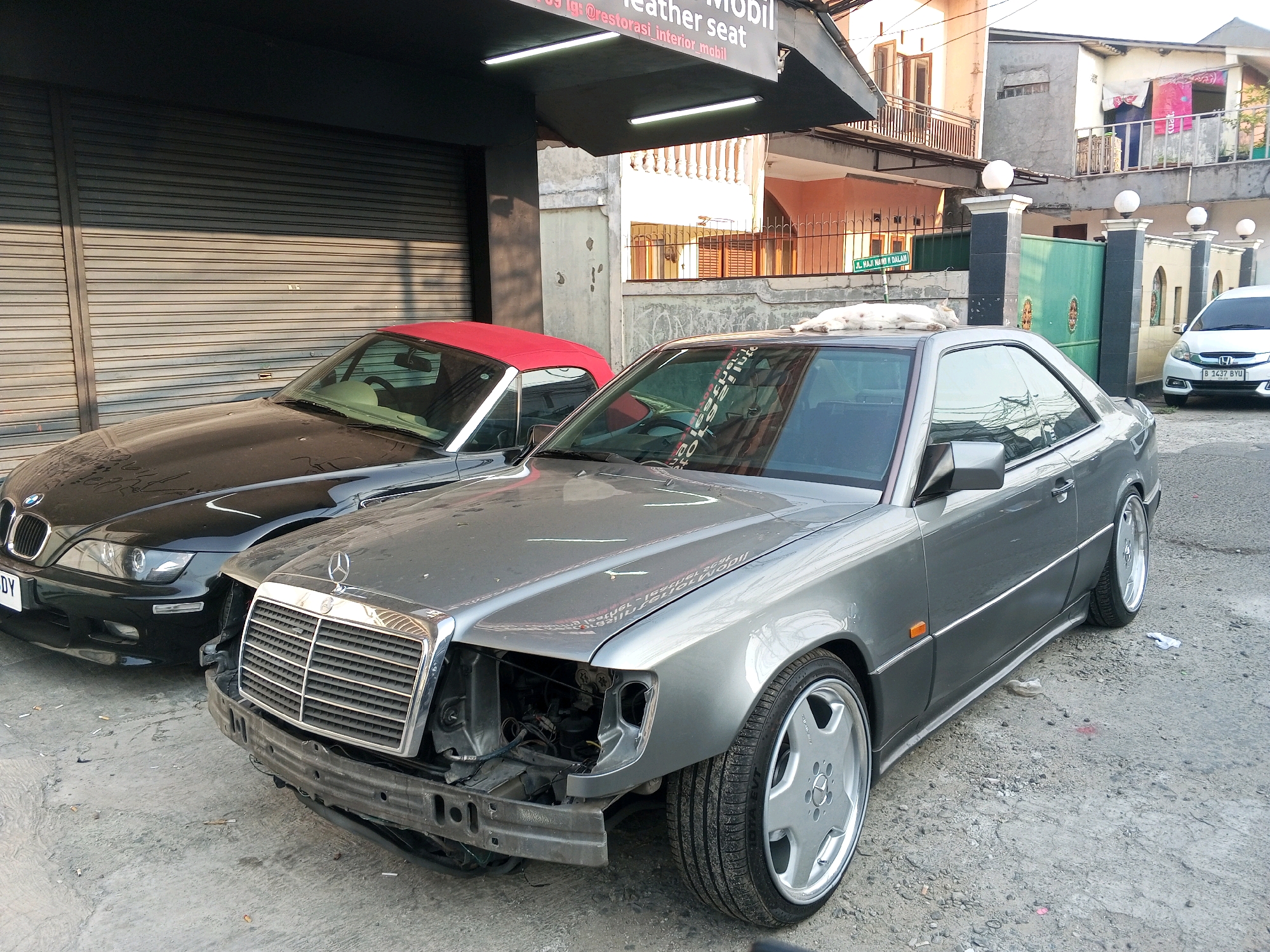 In the foreground, a damaged grey Mercedes-Benz coupe is parked, missing its front bumper and one headlight. The car has a distinctive silver multi-spoke wheel. To its left, a black convertible BMW with a red soft top is parked. Behind the cars, a closed grey roller door of a building is visible. Above the roller door, a black awning with fluorescent lights is installed. A white sign is attached to the awning, displaying text in Indonesian that appears to be an advertisement for "restorasi_interior_mobil" (car interior restoration). In the background, residential buildings with balconies and windows are visible. A white cat is lying on the roof of the black BMW. A white Honda Brio is parked to the right of the Mercedes-Benz. A green metal gate and a wall with decorative pillars are also in the background. A street sign with Indonesian text is attached to the wall. The scene appears to be an outdoor parking area in a South Jakarta, Indonesia neighborhood.