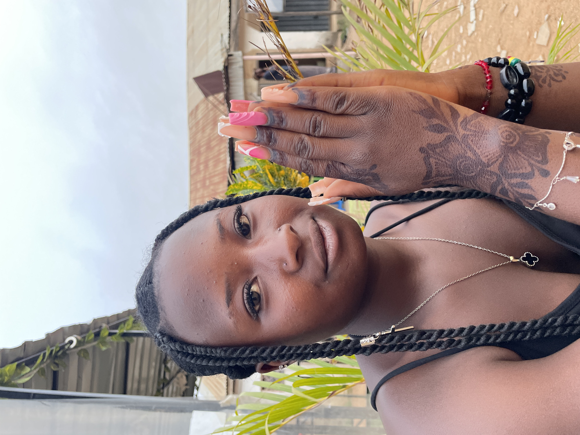 A close-up, slightly rotated shot shows a Black woman with braided hair and a nose piercing. She's wearing a black top and a silver necklace with a clover pendant. Her hands, adorned with intricate henna tattoos, are folded together, displaying long, decorative nails. One wrist has a black beaded bracelet with red and green accents. The background includes a building with a reddish-brown roof and some greenery, possibly palm leaves. The sky is overcast. The location is Jos, Nigeria.