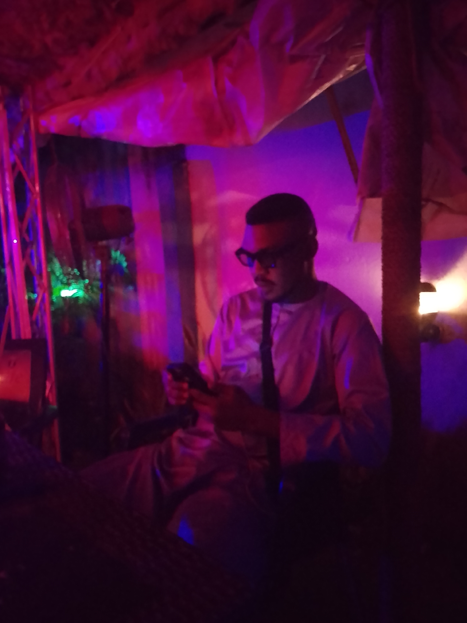 A dimly lit interior space, possibly a bar or lounge, in the city of Unknown, Unknown, features a man seated and looking at his mobile phone. The lighting is predominantly red and purple, with some green light visible in the background. The man is wearing dark sunglasses and a light-colored long-sleeved shirt. A vertical post is to his right, illuminated by a light. The ceiling appears to be draped with fabric. The environment is dark and the details are partially obscured due to the low lighting.
