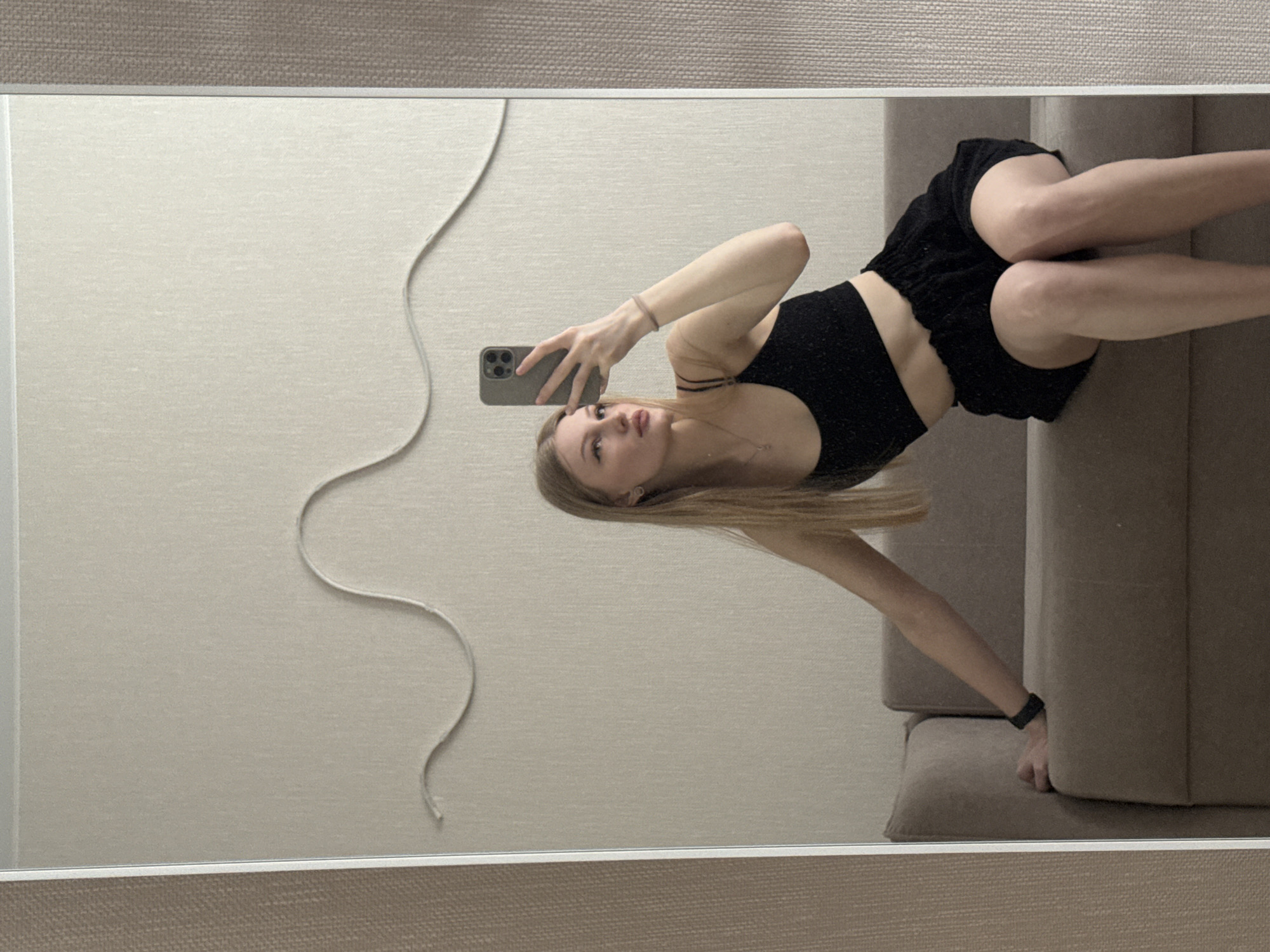 A young woman with blonde hair takes a selfie in a mirror. She is wearing a black crop top and black shorts. She is standing in what appears to be a room in a residential apartment. The wall is a plain beige color, and there is a white cord hanging down. She is leaning on a light brown couch that is against the wall. The location is likely in Perm, Russia.