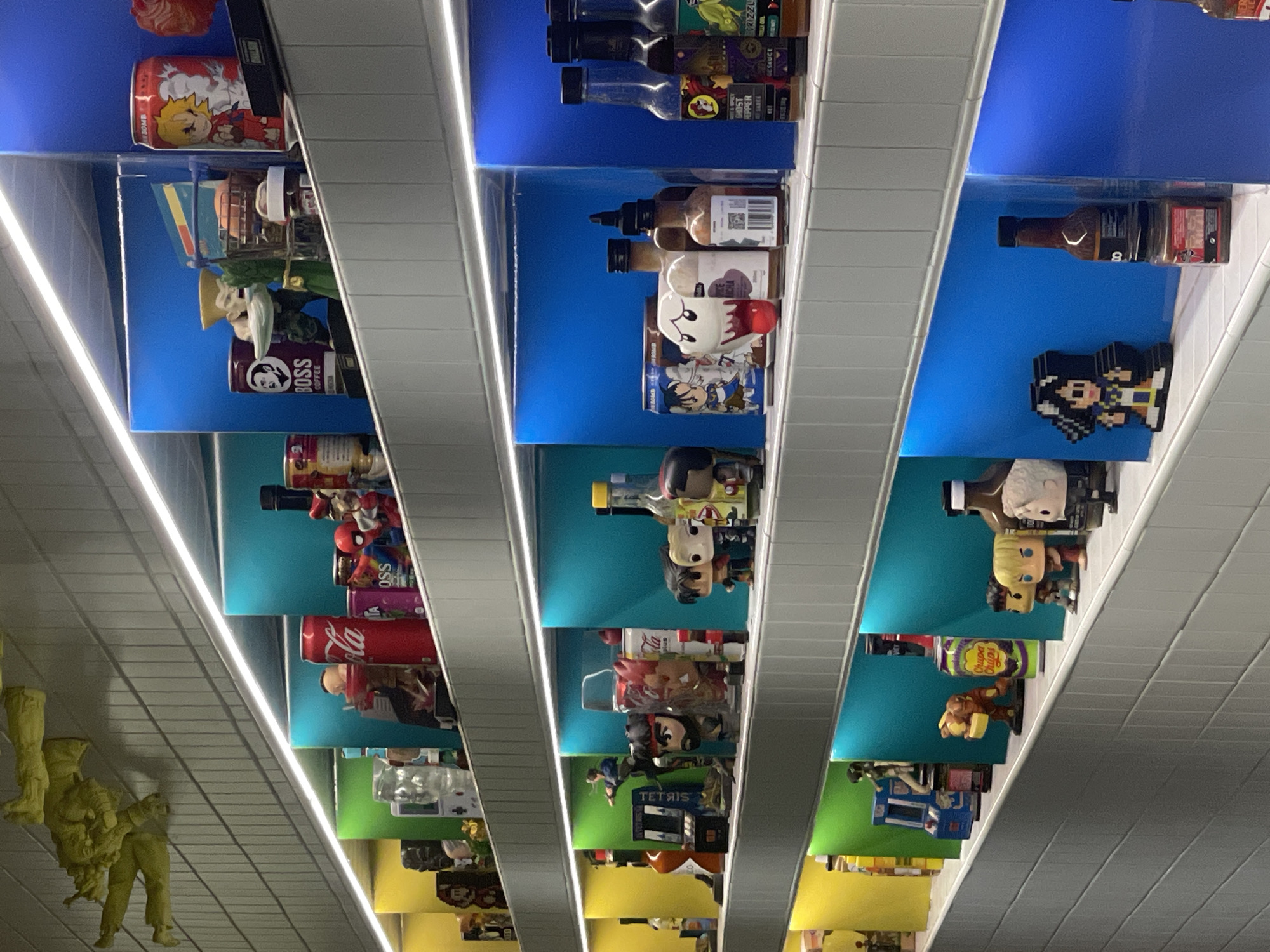 This is a well-lit, eye-level, medium shot of a display shelf featuring various collectible items and bottles. The shelves are angled, with each section illuminated by a colored light strip beneath it, creating distinct zones of yellow, green, and blue.
On the top shelf, a yellow, sculpted figure appears to be falling or in motion. Below this, the first visible section contains a red Coca-Cola can, a purple can with "BOSS COFFEE" text and a black silhouette of a man's head, a small Spiderman figurine, and other assorted figures and cans.
The next shelf down shows a variety of bottles and figures. Notably, there is a bottle with a yellow cap, a clear glass bottle, and several bottles of hot sauce with colorful labels. Figurines of characters from video games, such as Street Fighter, are prominently displayed.
Further down, more bottles and figurines are visible, including a Chupa Chups branded can, and characters that appear to be from anime or video games. The bottom shelf shows more hot sauce bottles and pixelated character figures.
The background consists of a light-colored, ridged wall. The overall impression is a curated collection of pop culture memorabilia and beverages. The scene does not provide any specific visual cues to pinpoint its location within Brisbane, Australia.