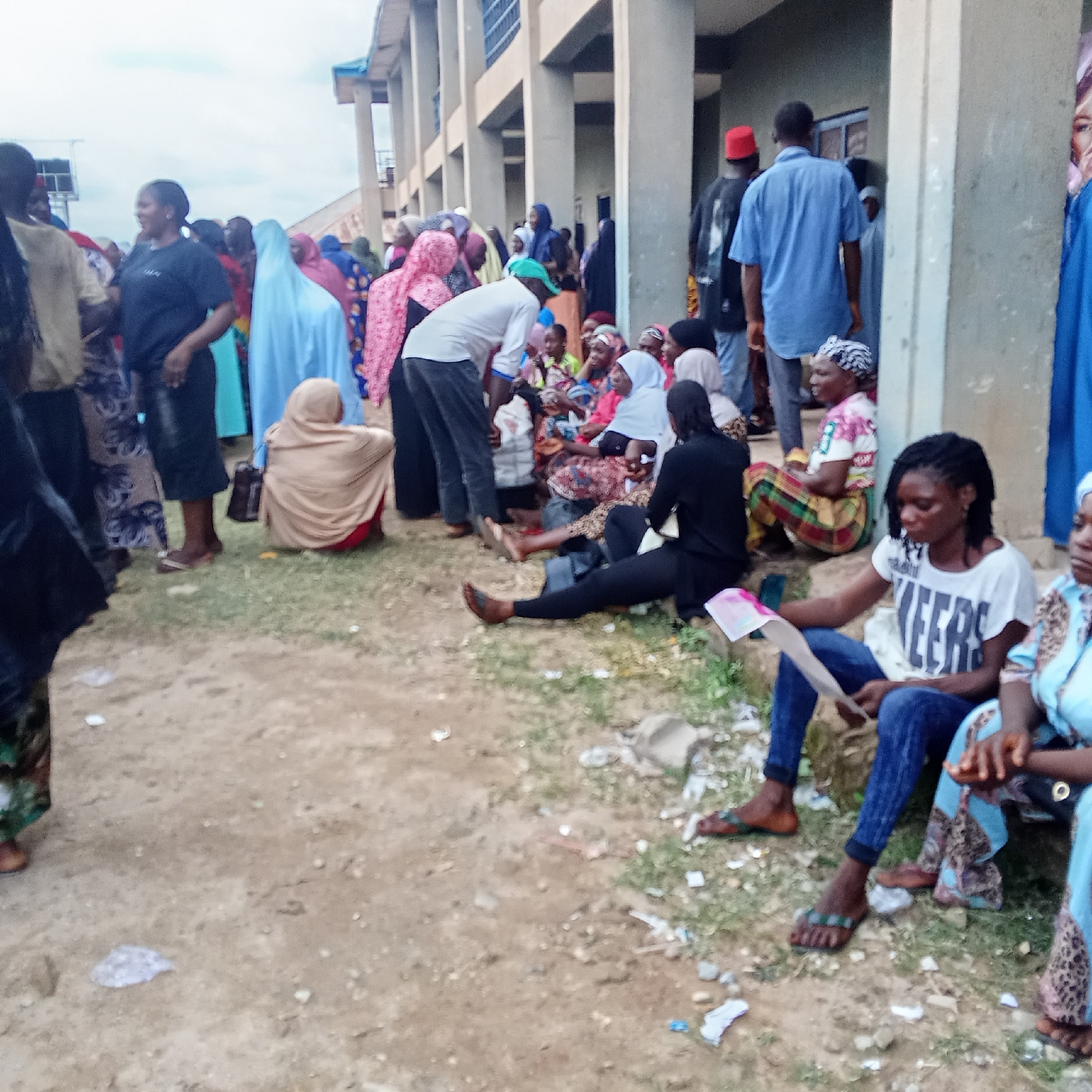 A gathering of people is observed near a building in Akwanga, Nigeria. The group includes women wearing headscarves, seated on the ground. One woman is holding a paper or document. Men are also present, some standing and some tending to those seated. The environment includes a building with columns and windows, and the ground is covered in grass and debris. The scene suggests a waiting or gathering area.