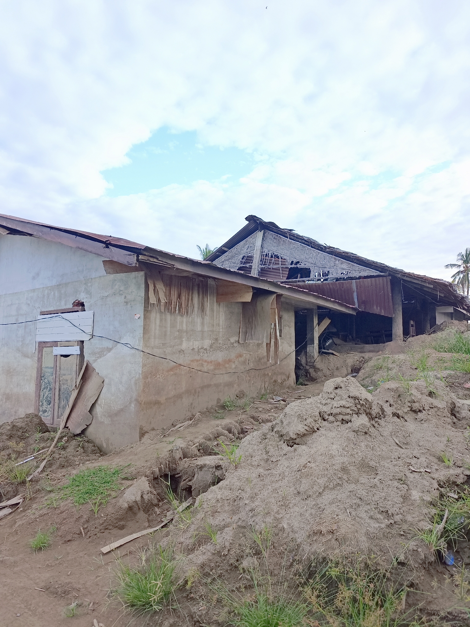 The image shows a damaged building in Meunasah Raya, Indonesia. The building's roof is severely damaged, with corrugated metal sheeting and wooden planks exposed. The walls of the building appear to be made of concrete and are stained and weathered. There is debris and mud piled around the building, suggesting it has been affected by a natural disaster or significant damage. The surrounding environment appears to be overgrown with grass and vegetation, and there are mounds of dirt and soil. The sky is cloudy, with patches of blue visible. No people are visible in the image.